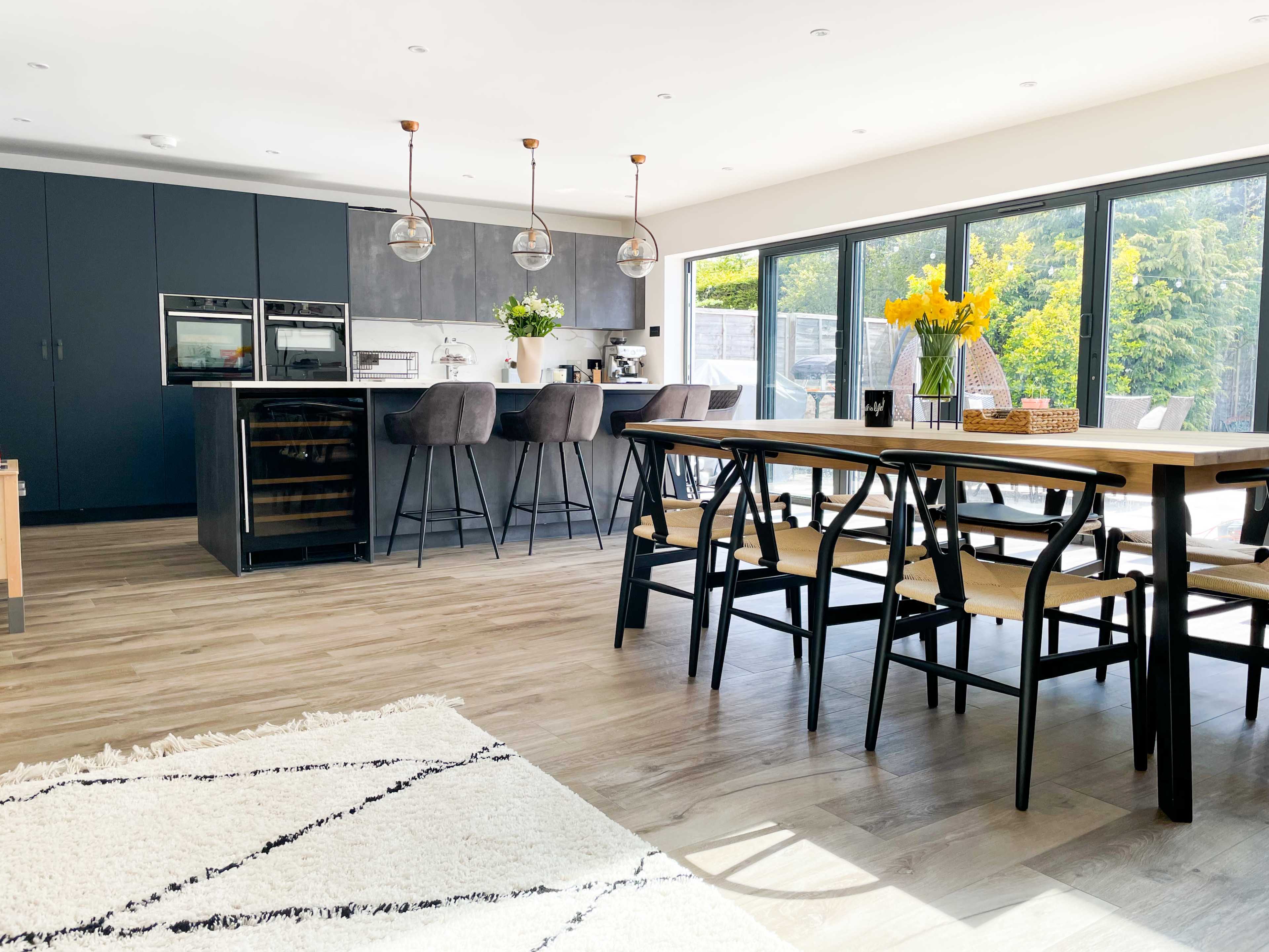 A modern kitchen and dining area features a large wooden table, dark cabinetry, and large windows overlooking a garden.