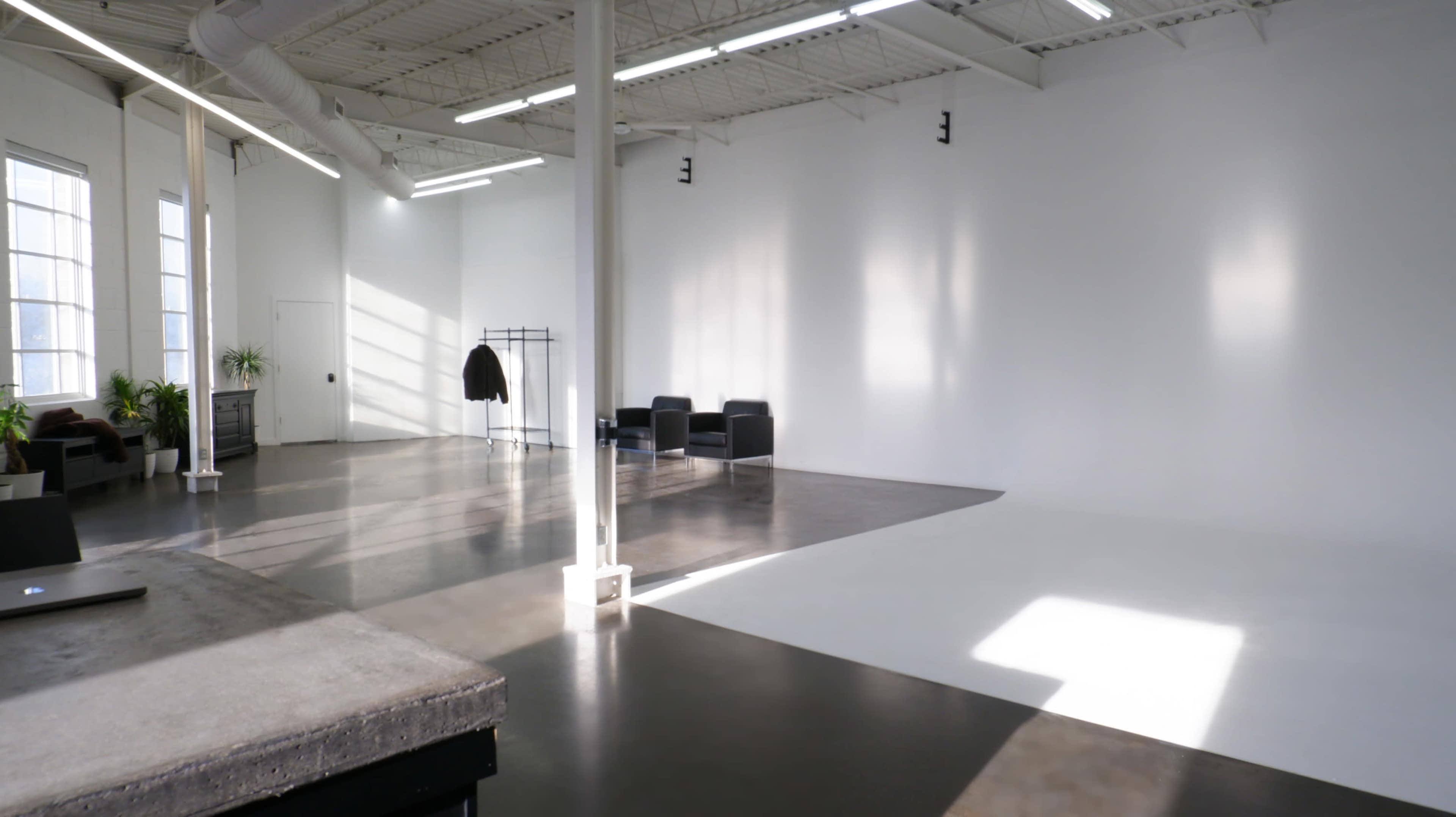 A spacious, bright studio with a polished concrete floor, a white backdrop, and minimalist furniture including a coat rack and two black chairs.