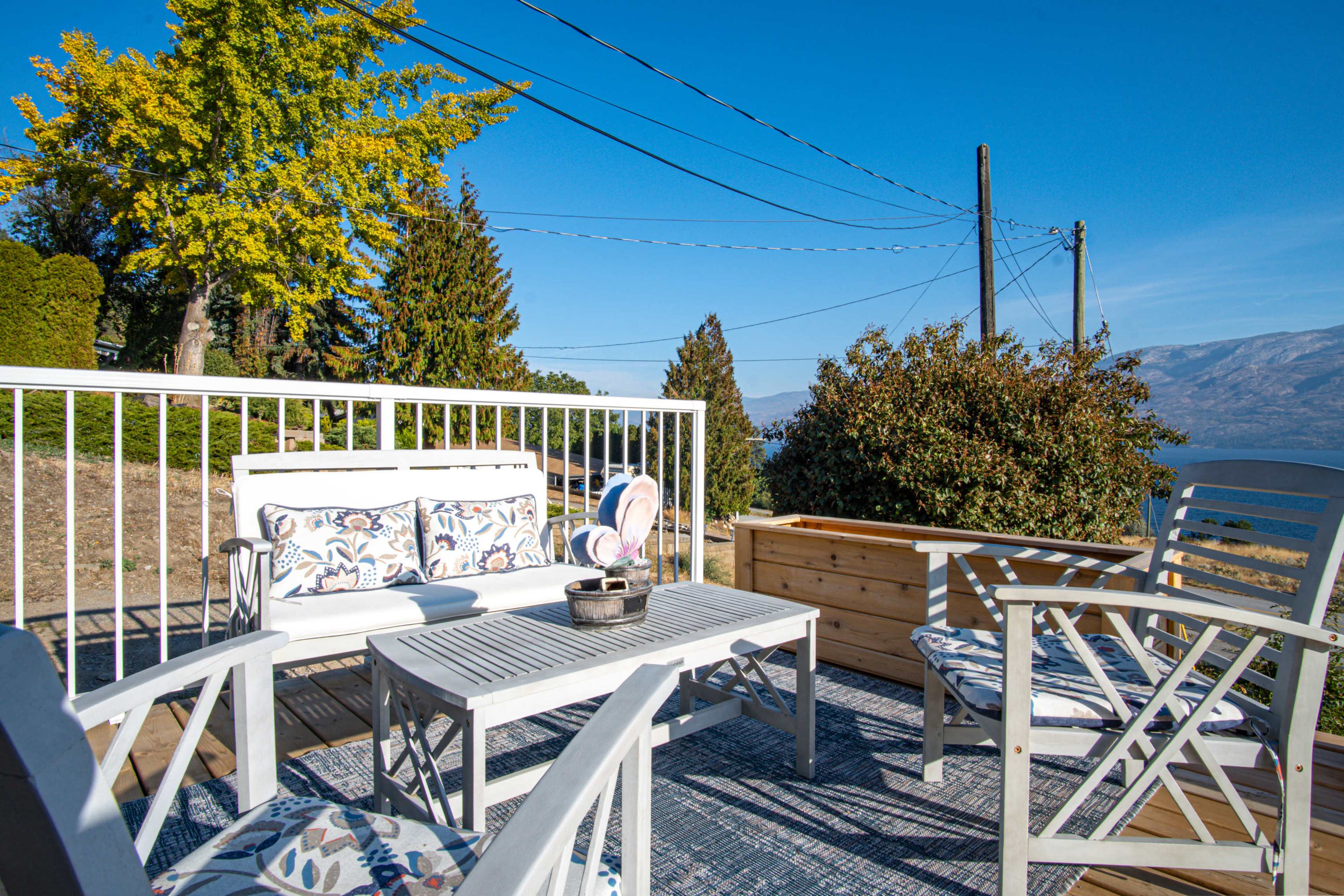 A sunny outdoor seating area features white furniture and a wooden planter, surrounded by trees and mountains under a clear blue sky.