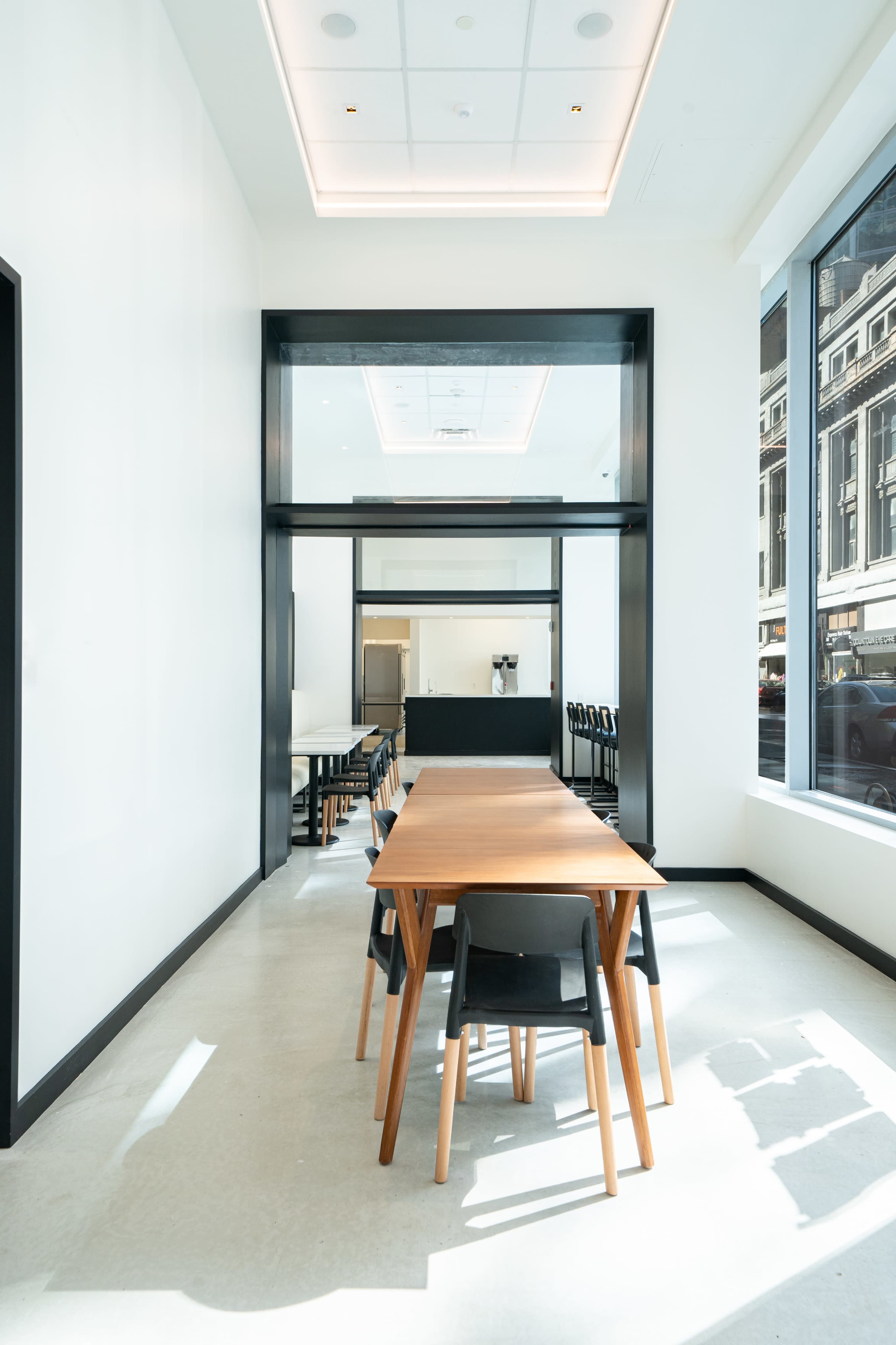 A wooden table with chairs is positioned in a bright, minimalist room with large windows and a modern design.