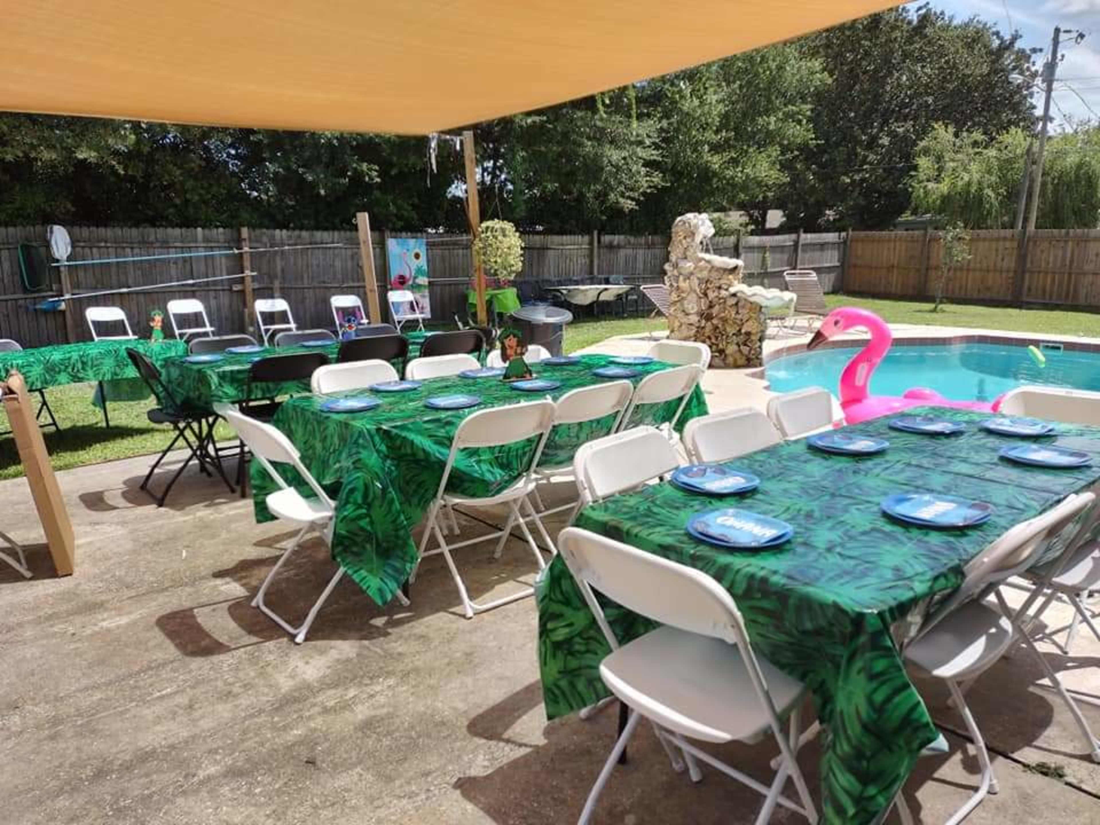 A backyard setup with multiple tables covered in leaf-patterned tablecloths, arranged around a pool area adorned with a pink flamingo and festive decorations.