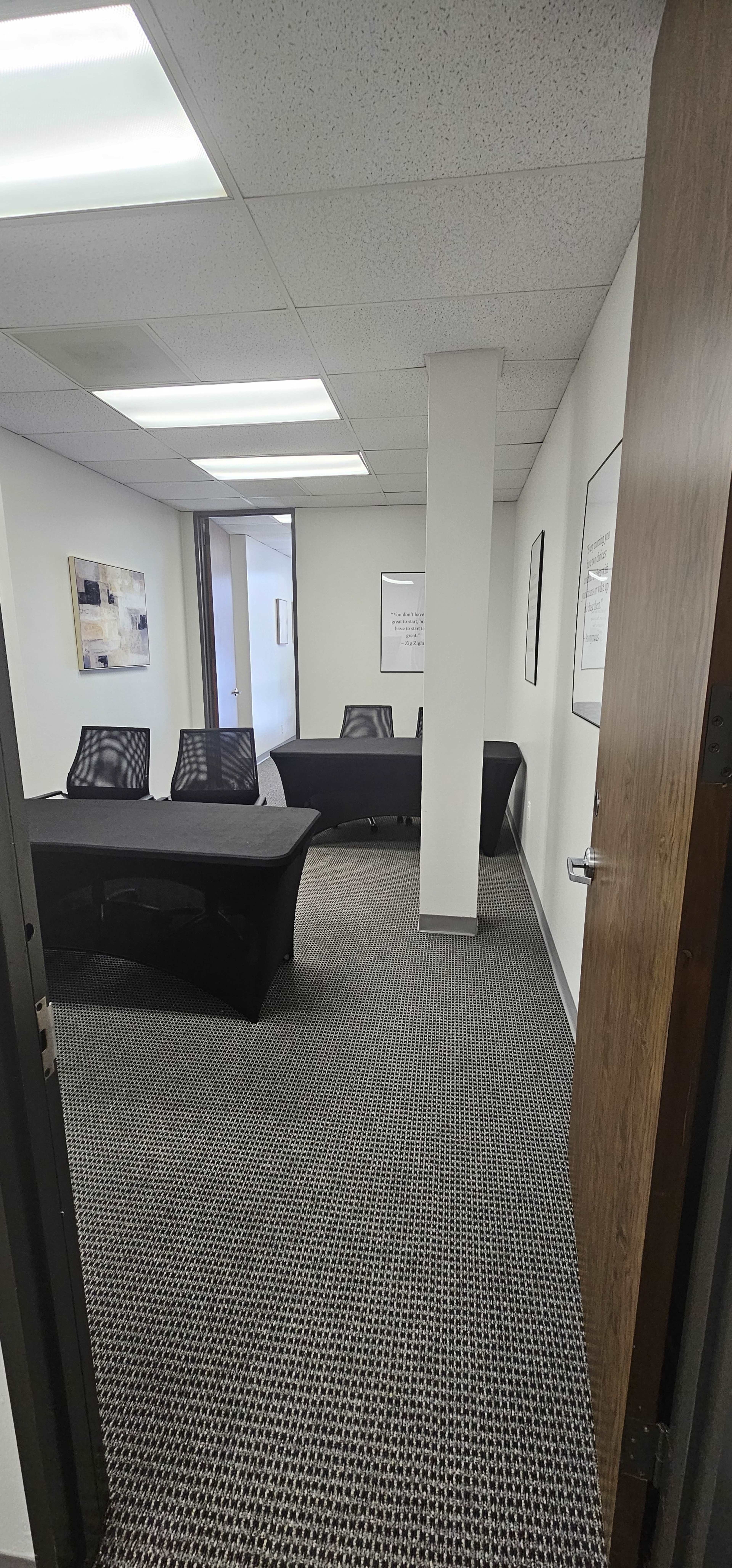 The image shows a conference room with a long black table and several black chairs arranged along its sides, set within a carpeted office space.