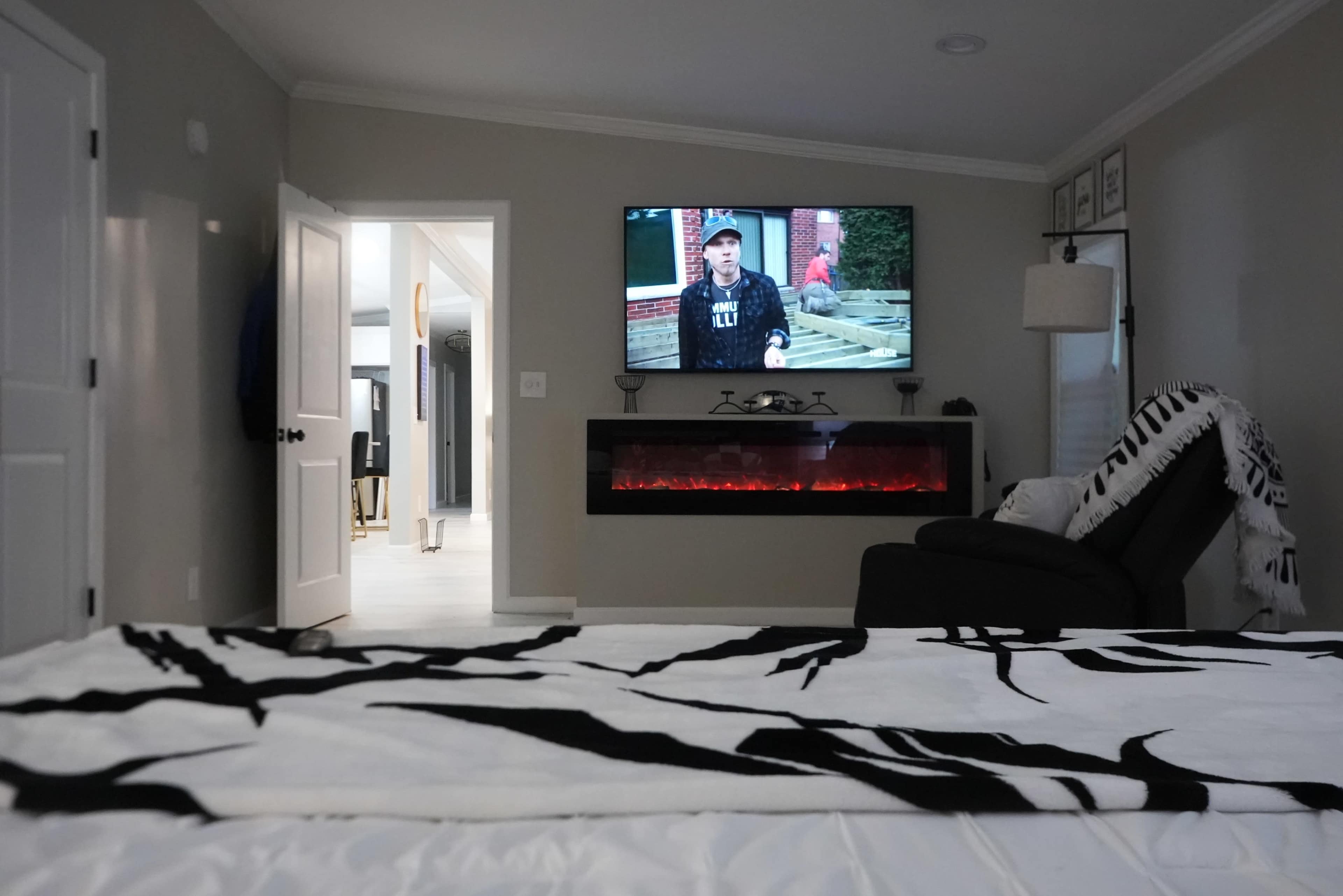 The image shows a cozy bedroom featuring a large flat-screen TV mounted on the wall, an electric fireplace beneath it with glowing embers, and a bed covered with a patterned blanket.