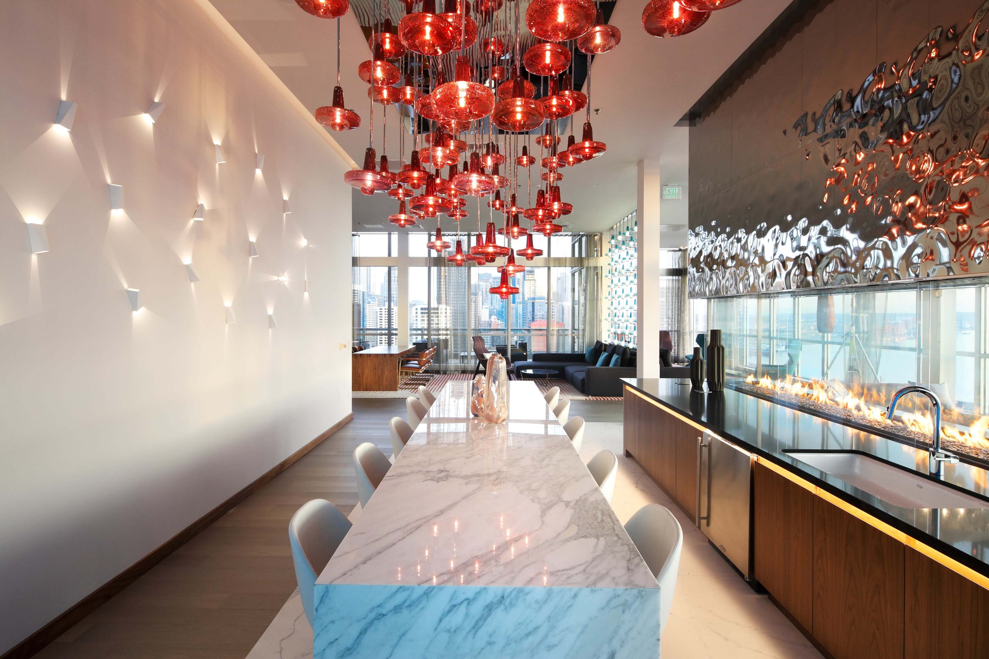 A modern kitchen and dining area featuring a long marble table with white chairs, accented by red pendant lights hanging above, and a sleek kitchen with a glass wall displaying a fire feature.