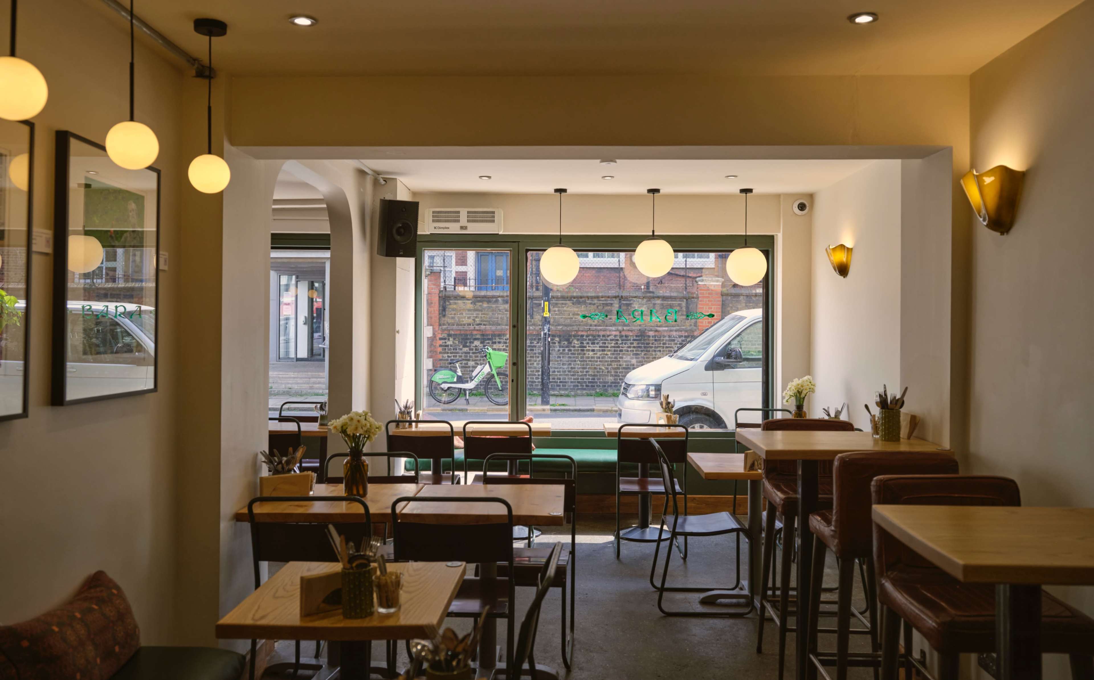 The image shows a bright, minimalist café interior with wooden tables and chairs, featuring large windows that offer a view of the street outside.