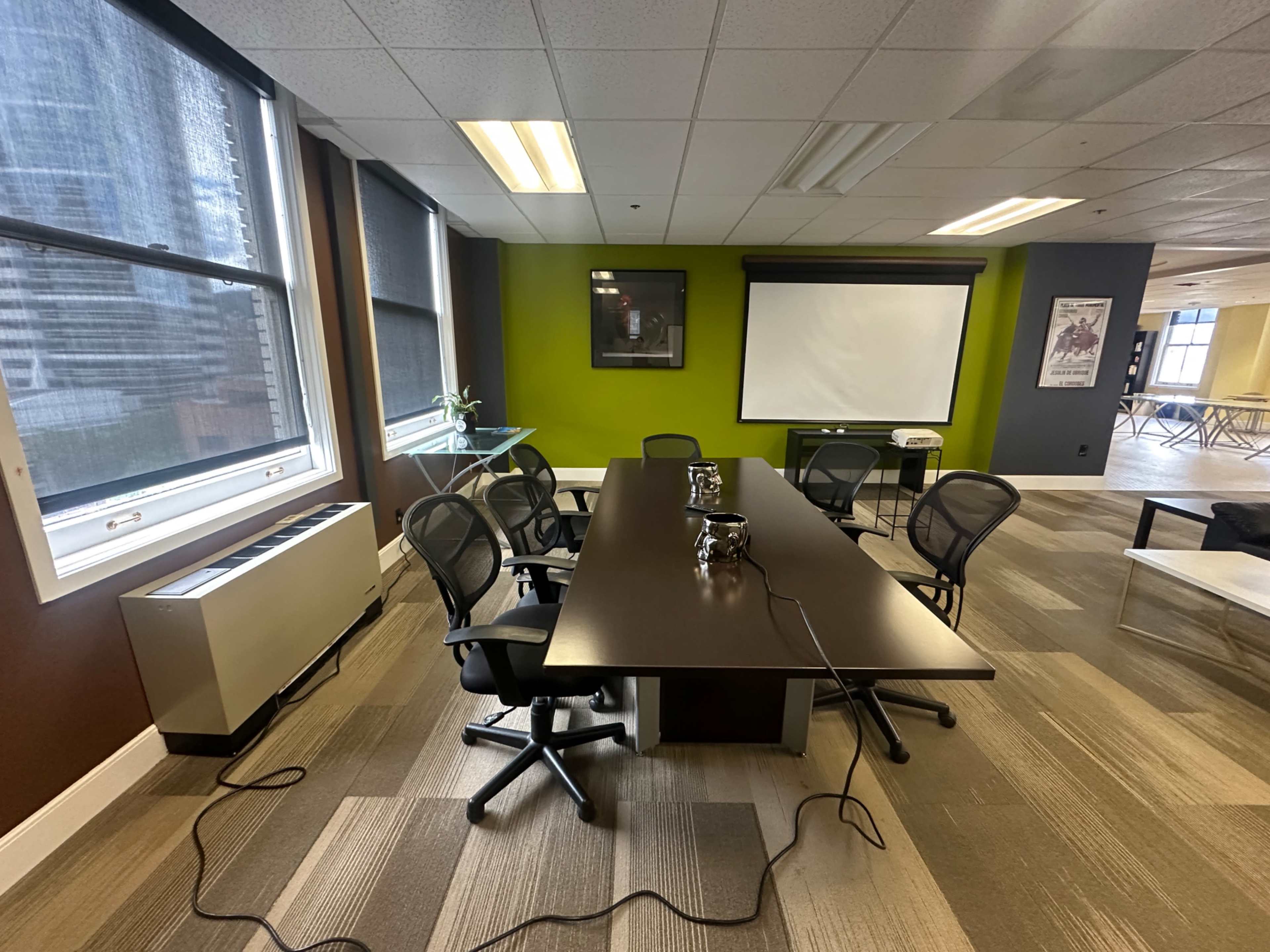 The image shows a conference room with a long table surrounded by several chairs, a projector screen on the wall, and large windows letting in natural light.