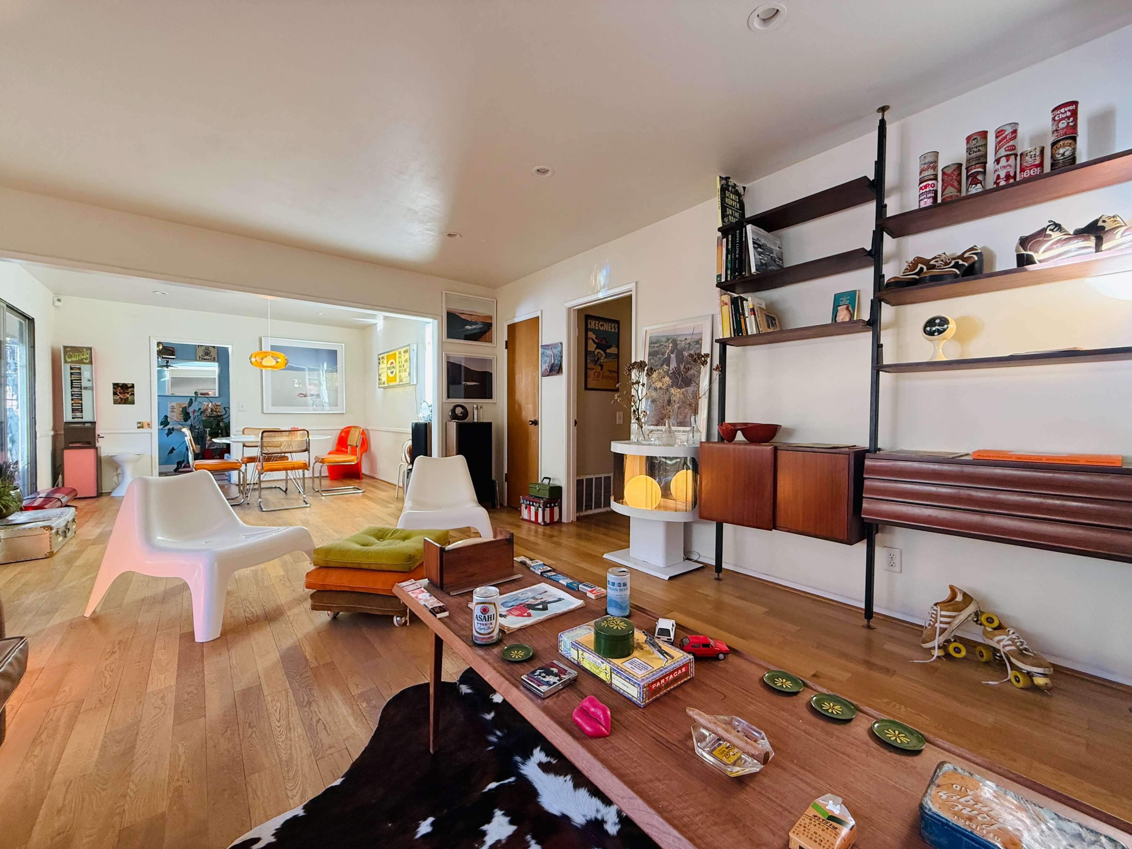 The image shows a mid-century modern living space with a mix of vintage furniture, including a table with snacks, a bright orange chair, and a bookshelf displaying decorative items.