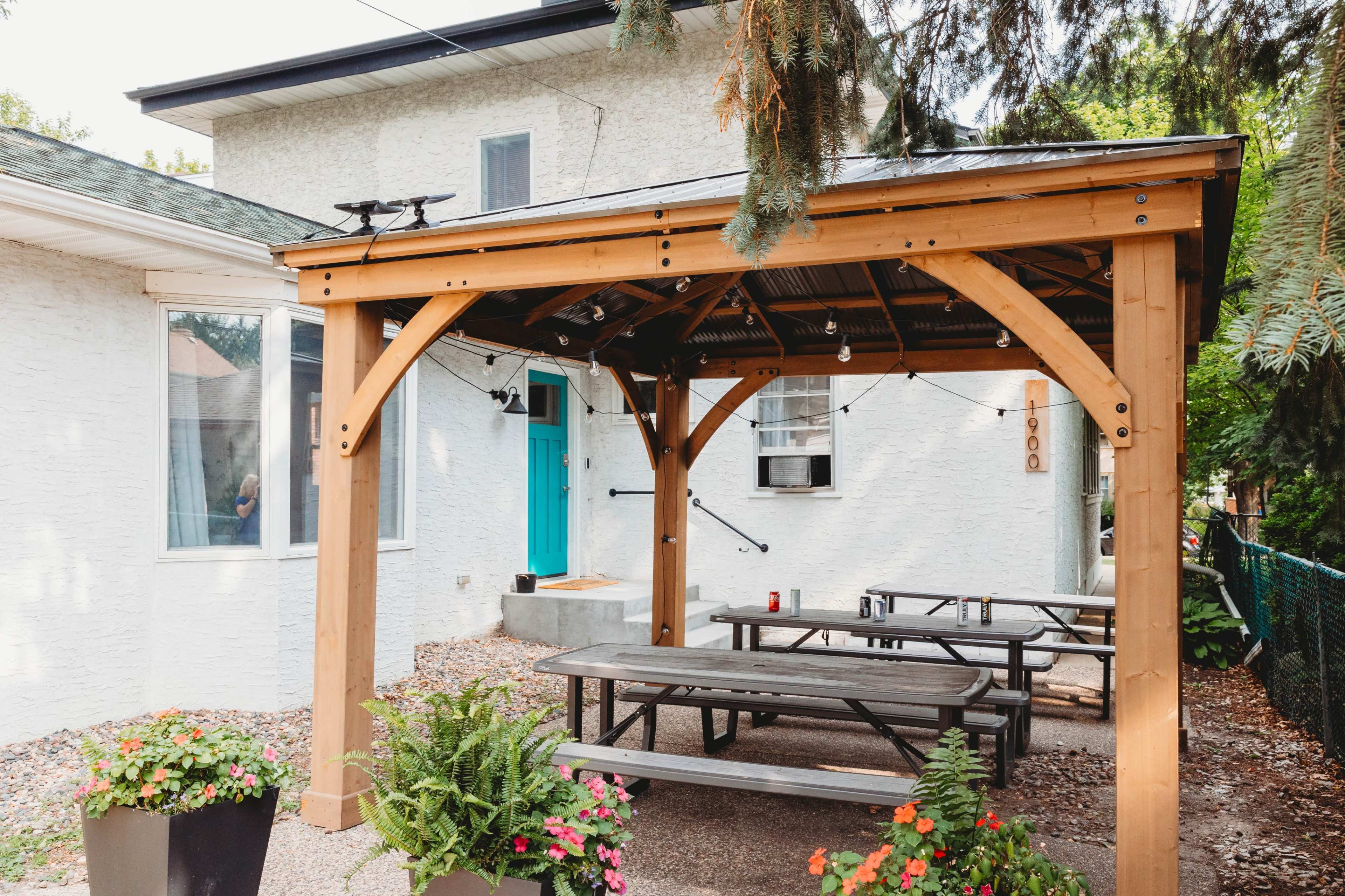 A wooden gazebo shelters several metal tables and chairs with potted plants and string lights, located in a gravel area next to a white house with a blue door.