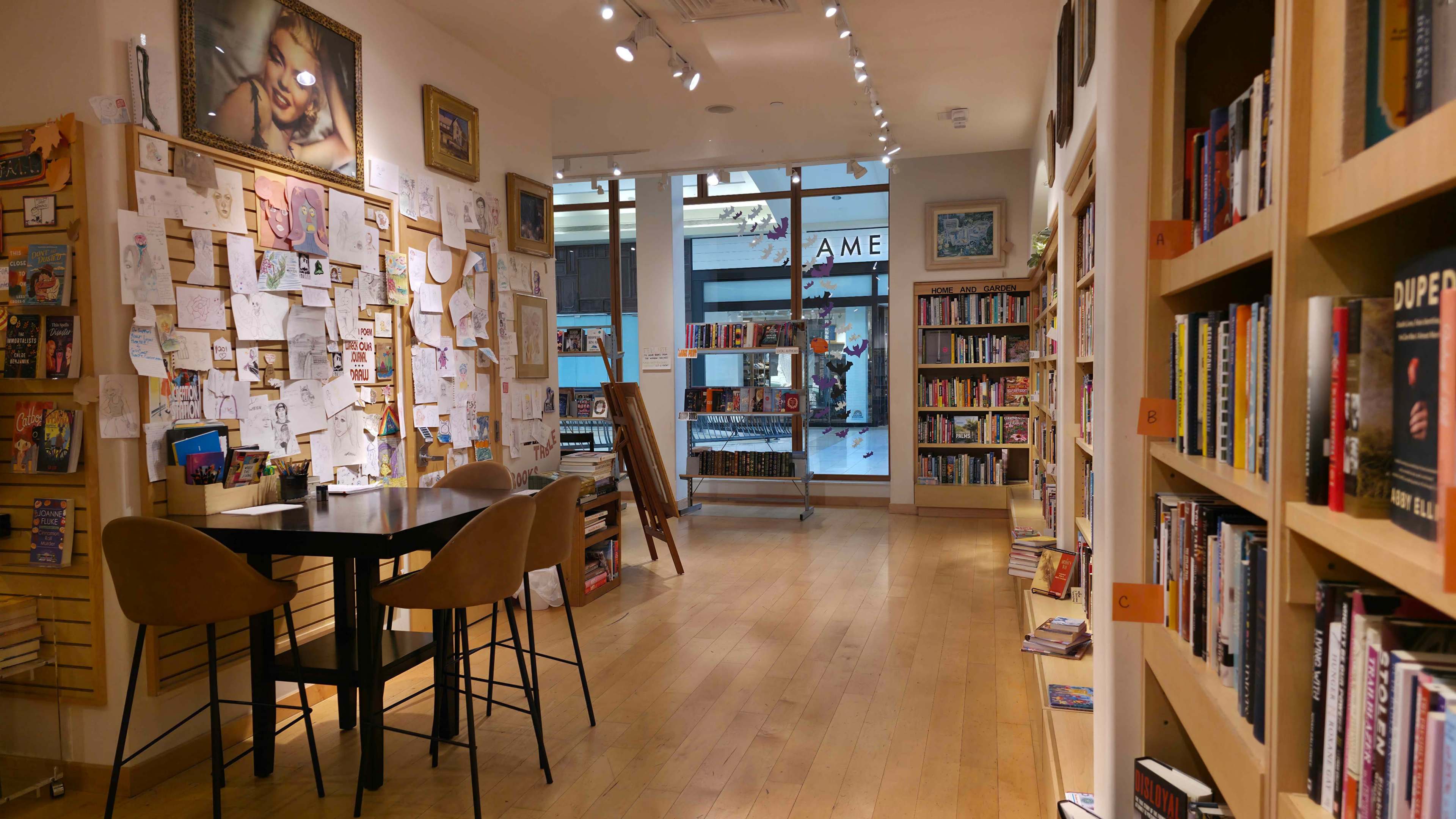 The image shows a cozy bookstore interior with wooden shelves filled with books, a table and chairs in the center, and a wall covered in various notes and artwork.