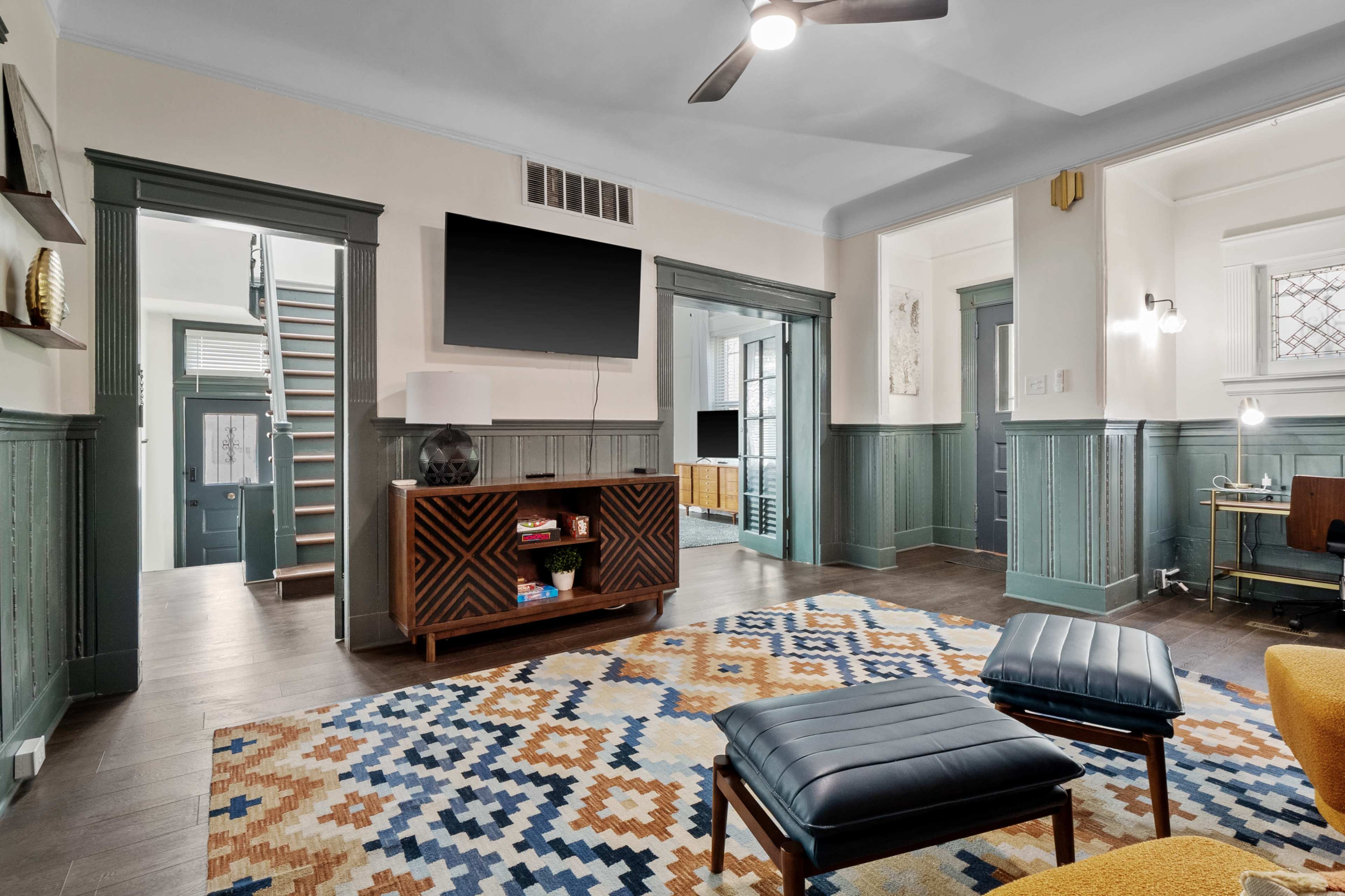 The room features a patterned rug, modern furniture, a mounted television, and a view into an adjacent room with a staircase.