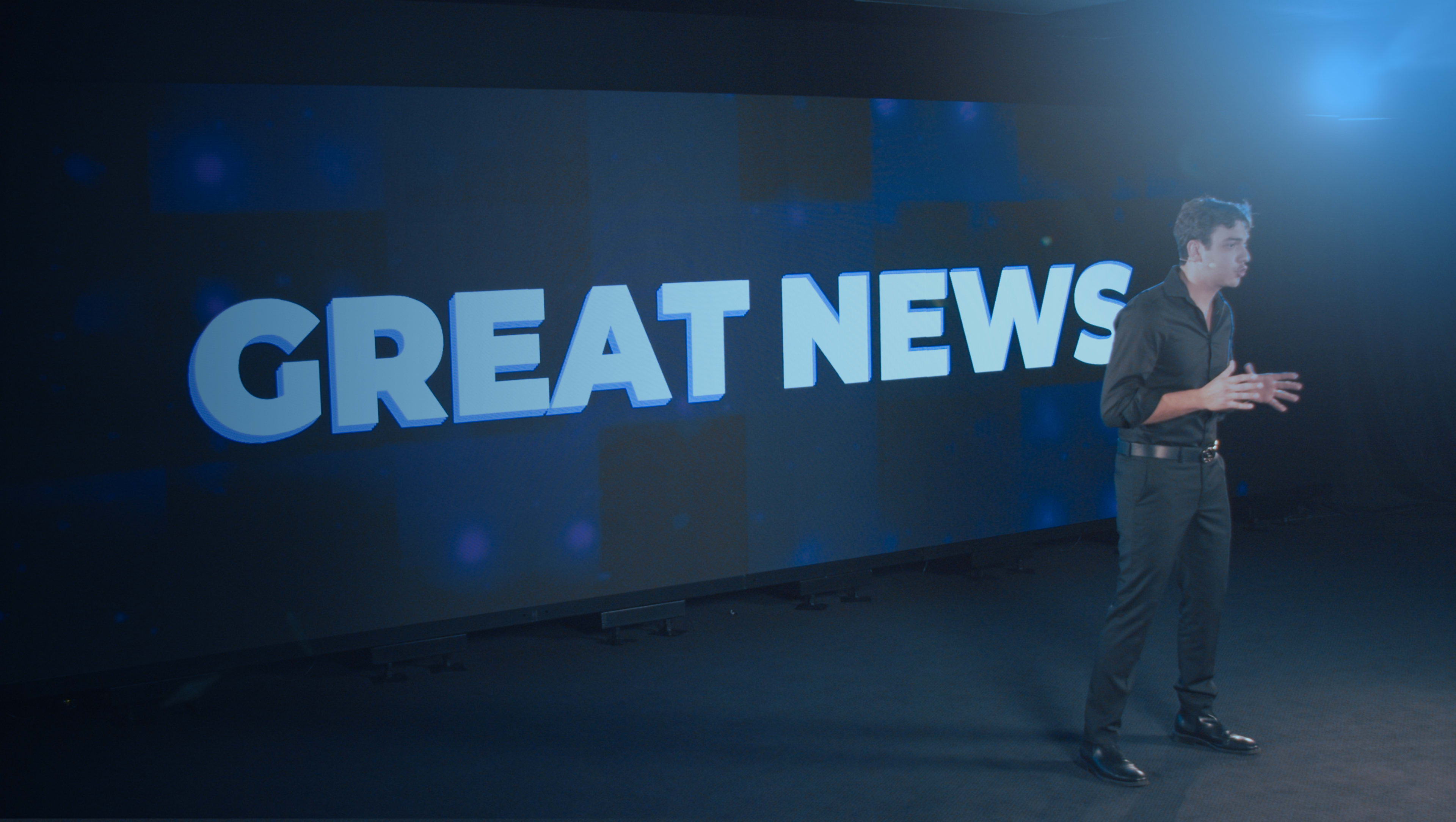 A person stands on a stage in front of a large screen displaying the text "GREAT NEWS."