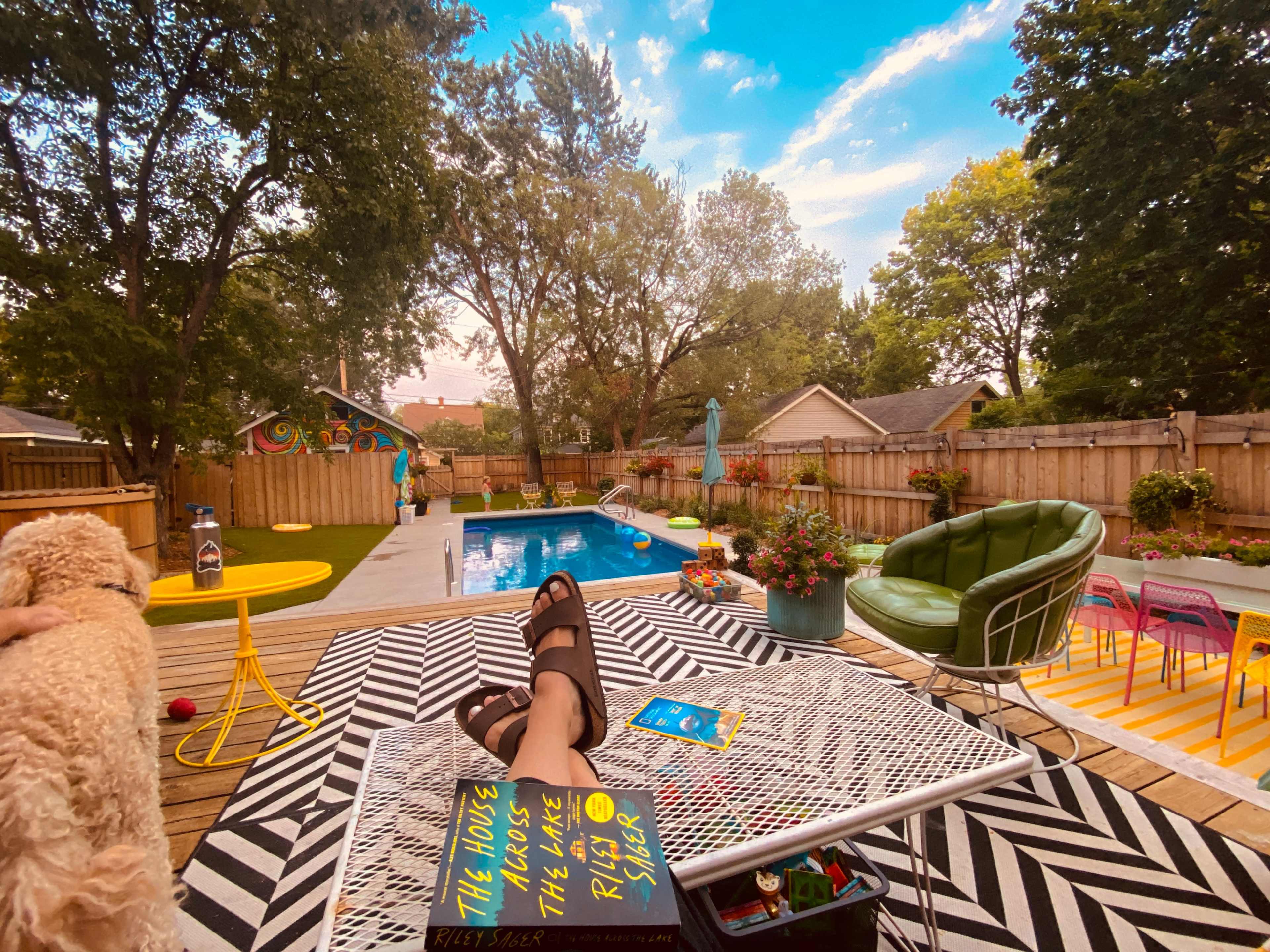 A backyard scene features a swimming pool surrounded by colorful lounge furniture and potted plants, with a person standing nearby and a small dog near the camera.