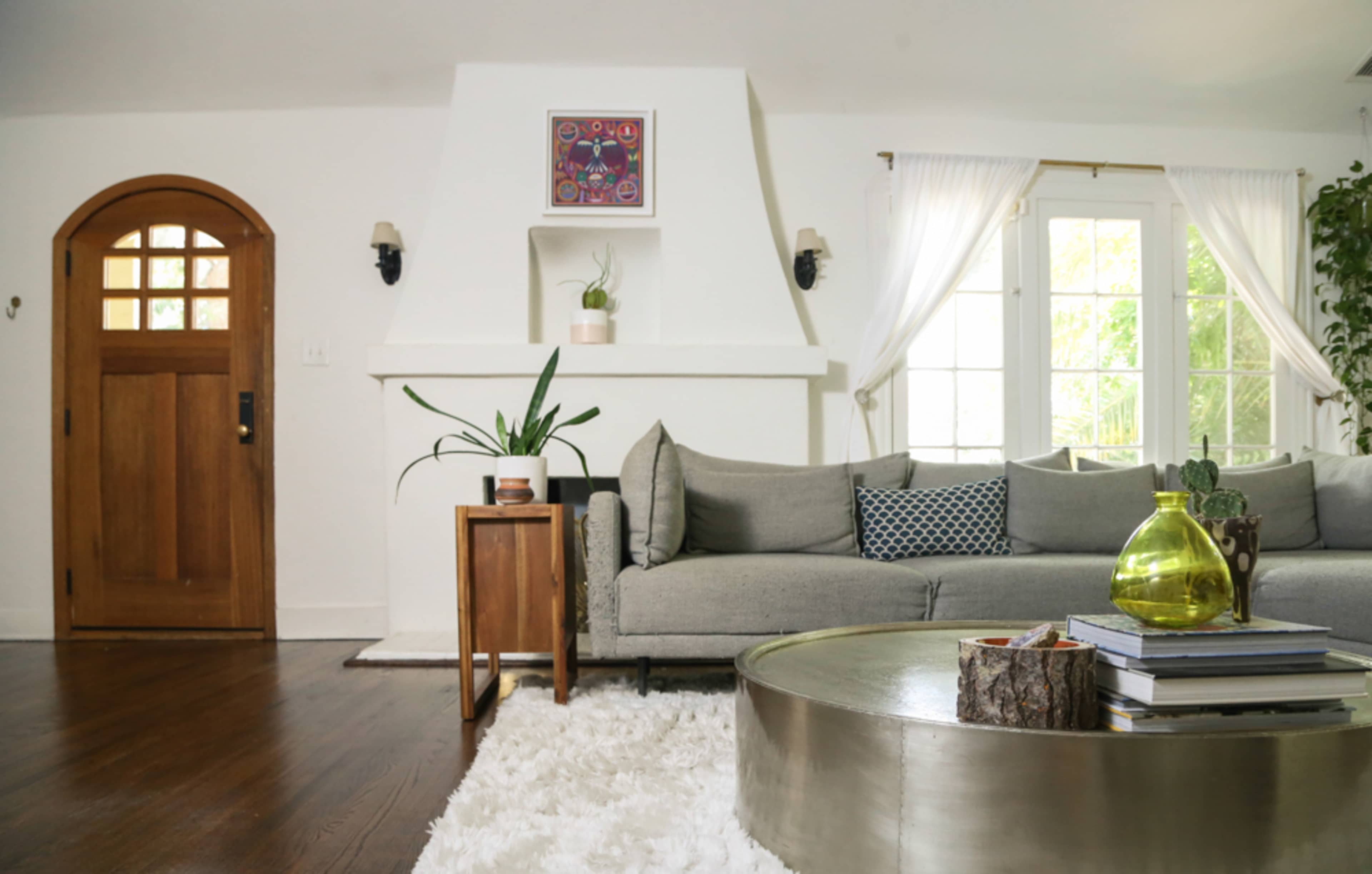 The image shows a modern living room with a gray sofa, a circular coffee table, a wooden front door, and a large window with sheer curtains.