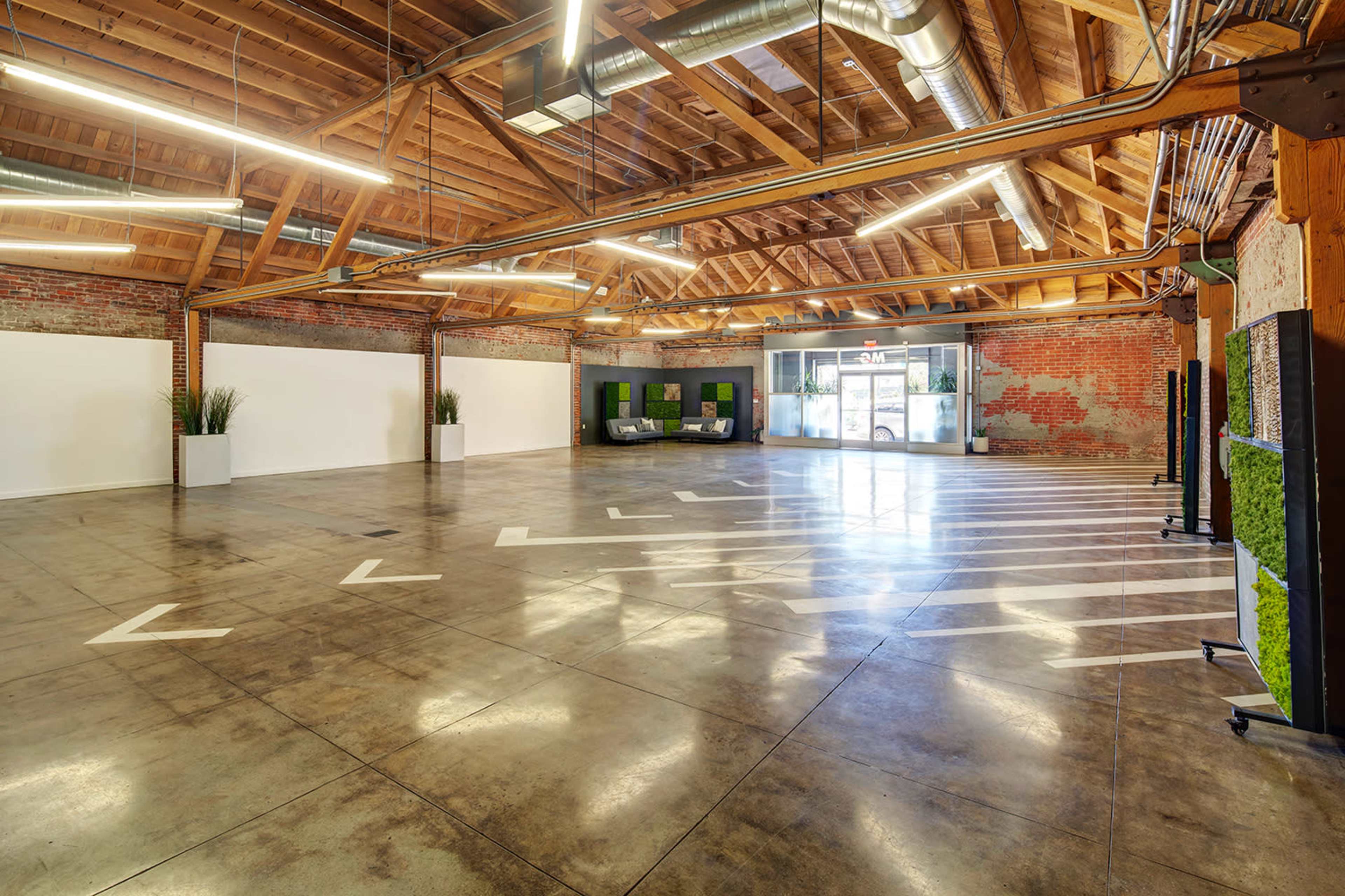 A spacious indoor area with a polished concrete floor, wooden beams, and large windows, featuring minimal furniture and greenery.