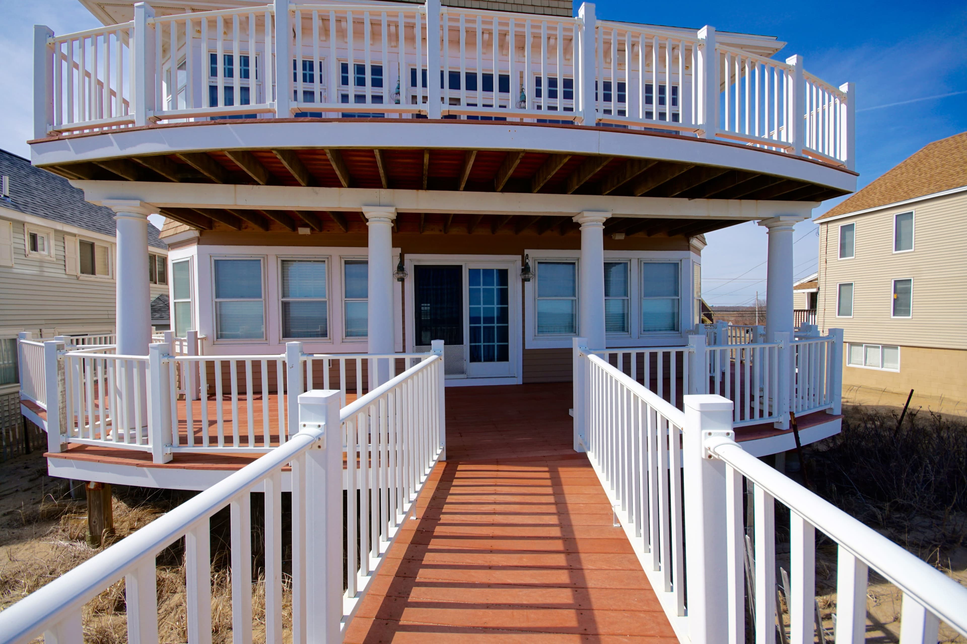 A two-story house with a large, elevated deck and white railings, accessed by a wooden walkway.
