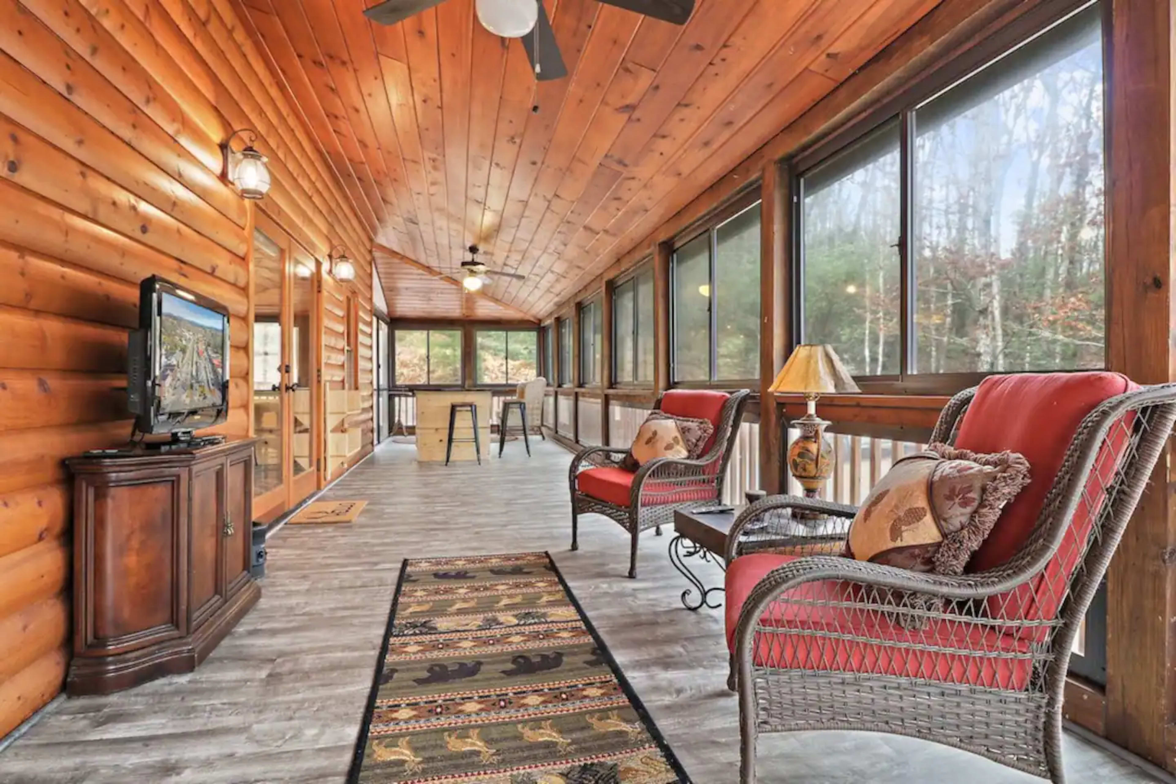 A wooden screened porch with a television, two wicker chairs, a small table, and large windows overlooking a forested area.
