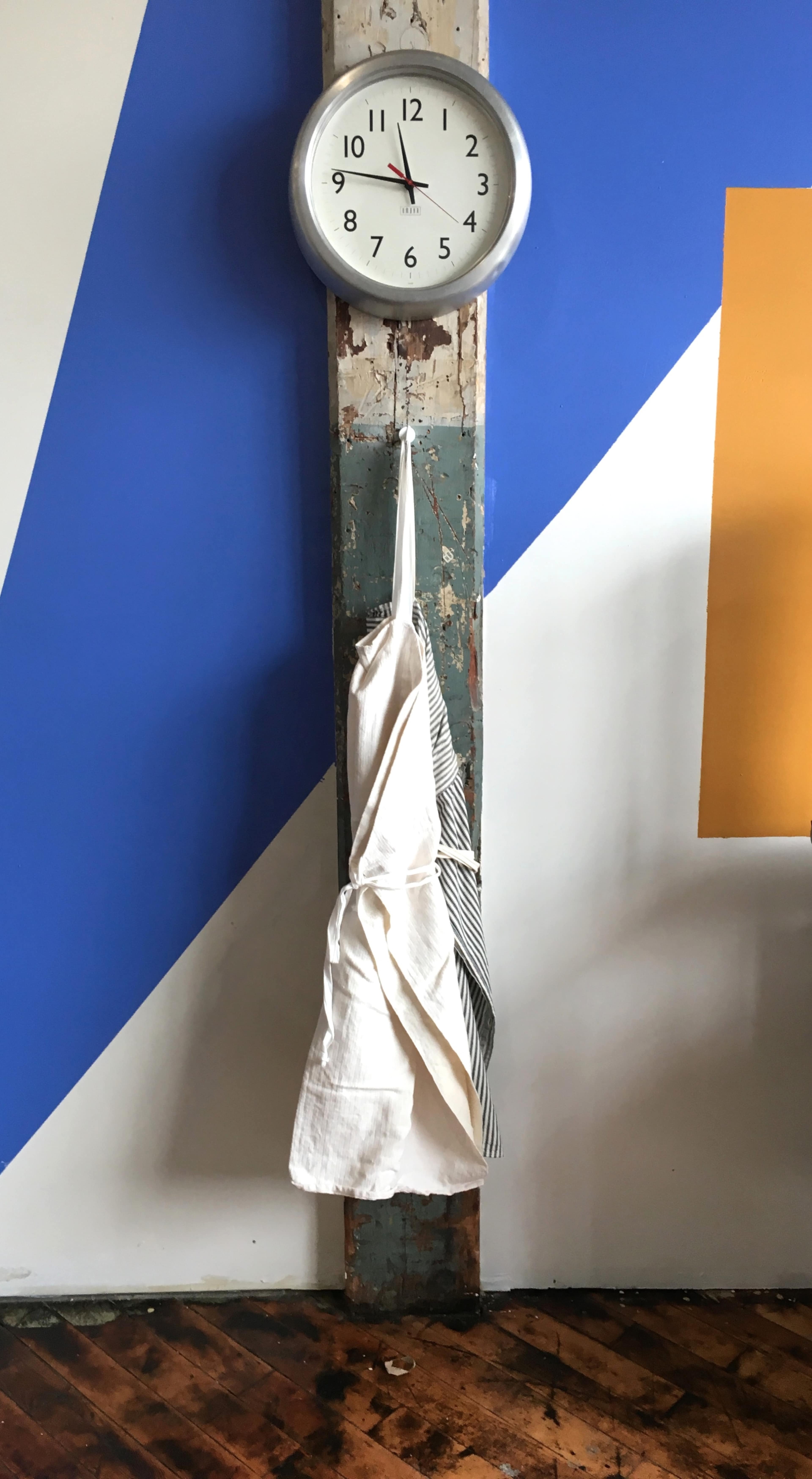 A white apron hangs on a post under a wall clock on a colorful geometric wall.