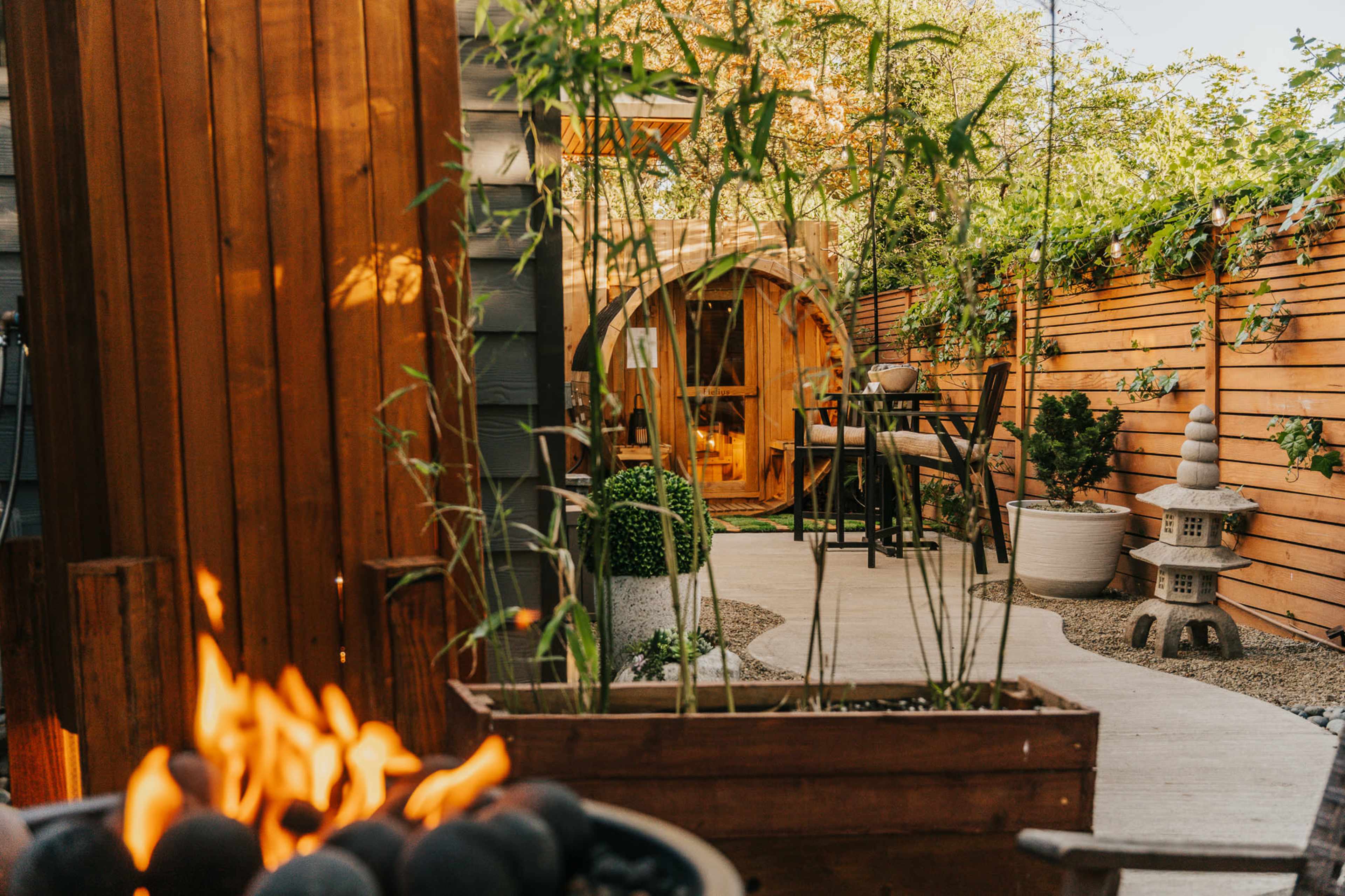 The image shows a landscaped garden featuring a circular wooden structure, a stone pathway, and a fire pit with flames in the foreground.