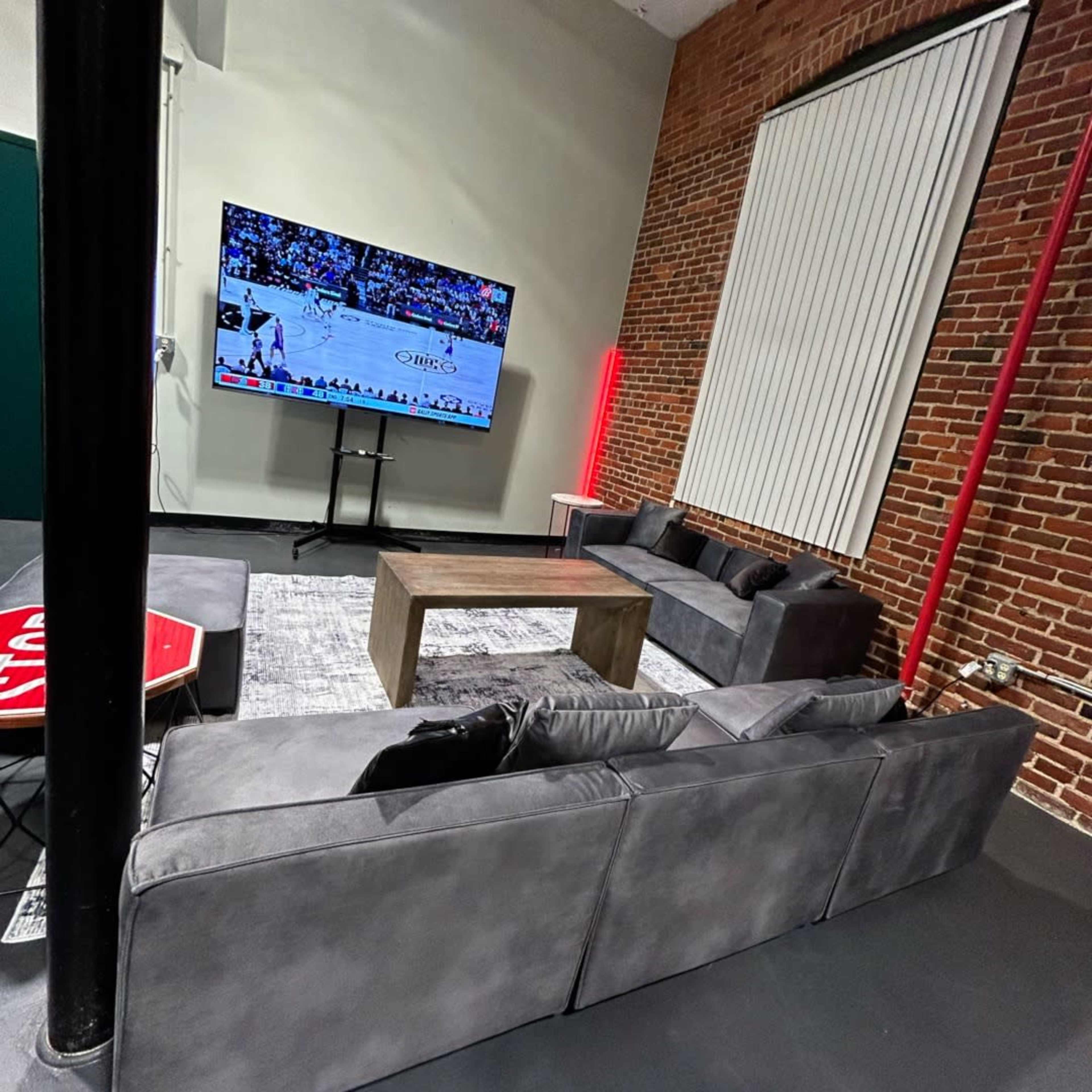 A modern living room with a large gray sectional sofa facing a wall-mounted TV that displays a basketball game, against a backdrop of exposed brick walls and a minimalist decor.
