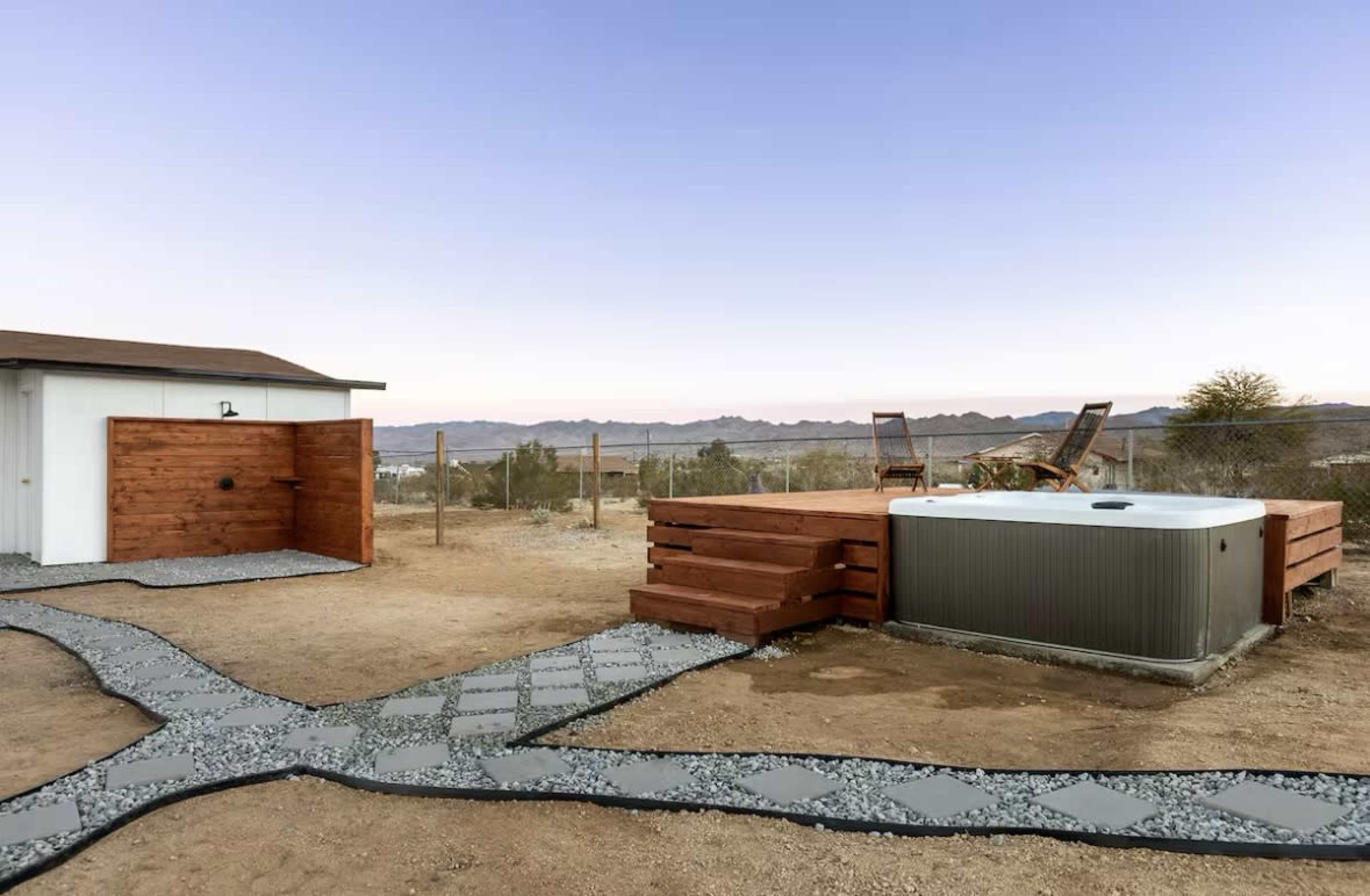 The image shows a desert landscape featuring a hot tub surrounded by wooden decking and a gravel pathway leading to a storage shed.