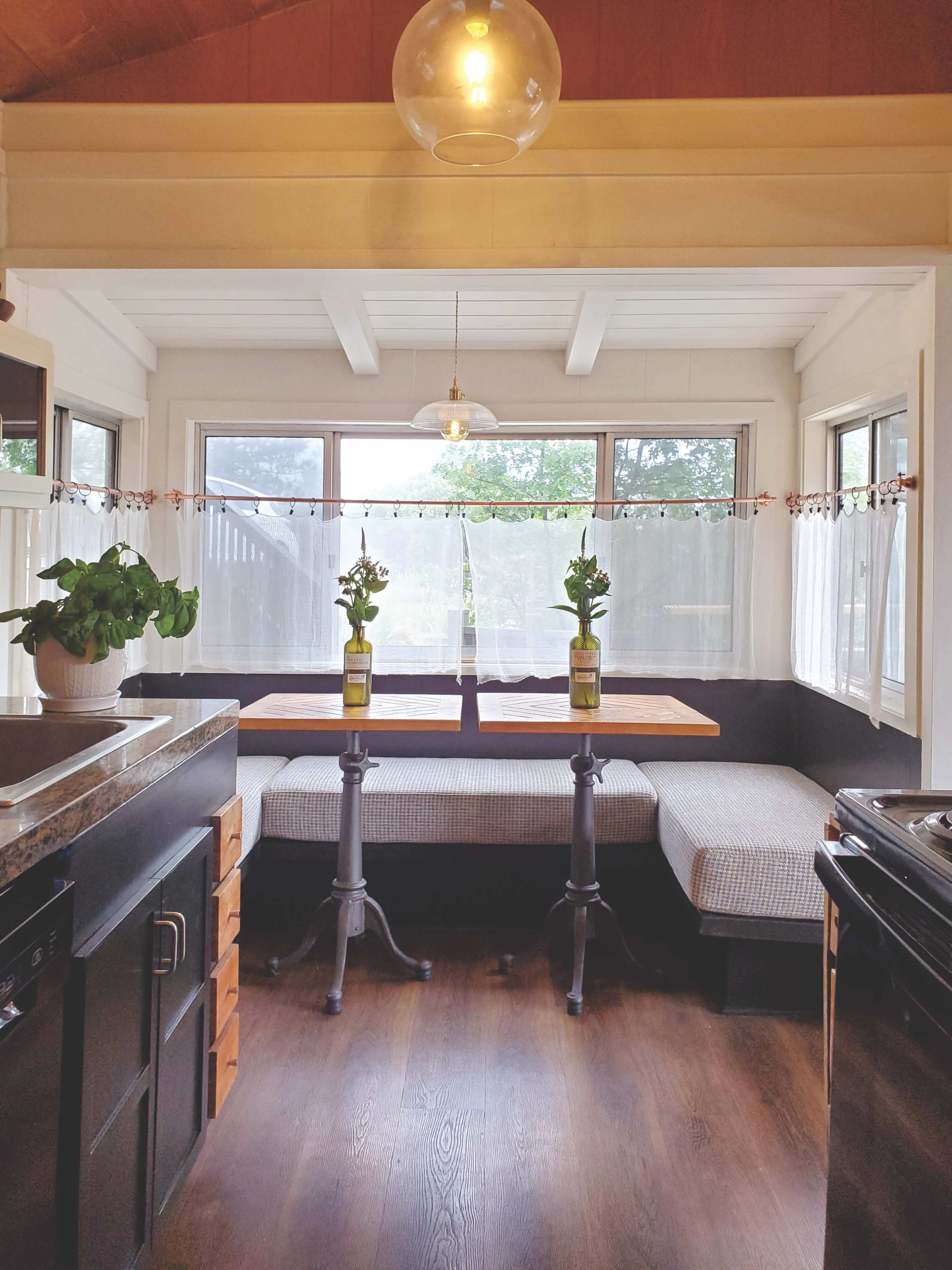 A sunlit dining area features a wooden table with two chairs on a cozy bench, surrounded by large windows and a potted plant.