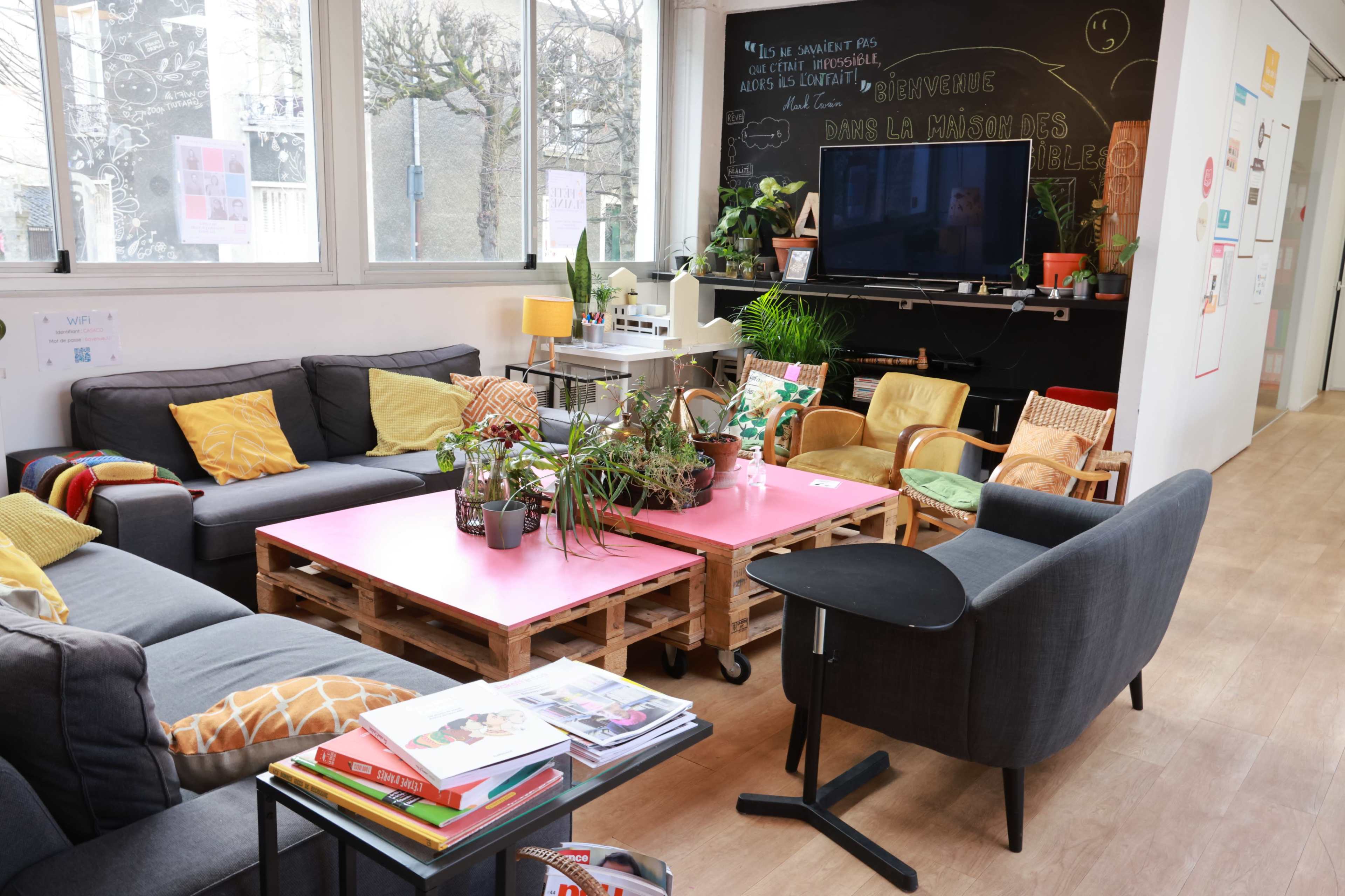 A cozy lounge area with grey sofas, colorful cushions, plants, and a pink coffee table made of pallets, set against a backdrop of large windows and a chalkboard wall.