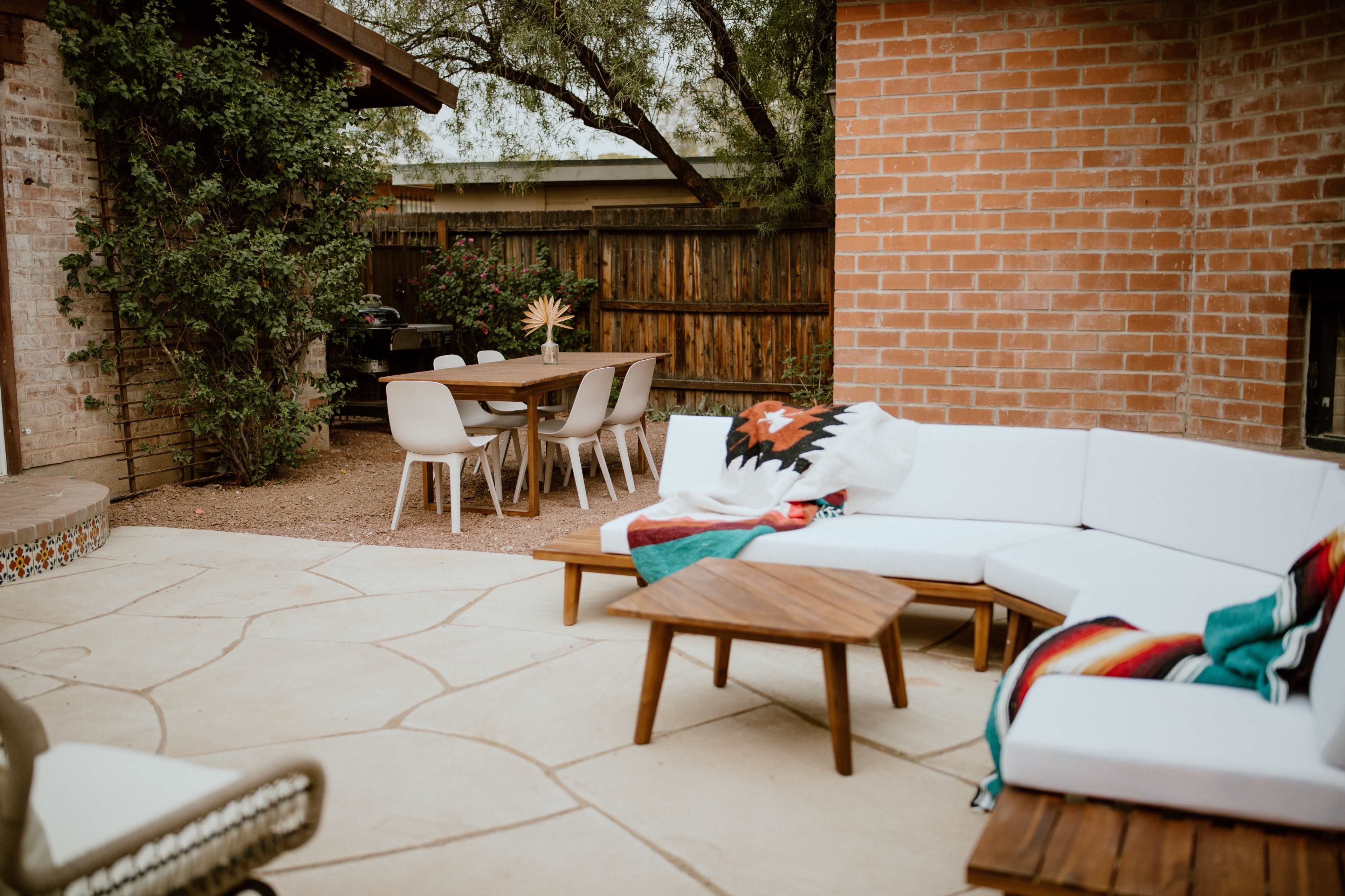A backyard patio area featuring a sectional sofa with a colorful blanket, a wooden coffee table, and a dining table with chairs under a wooden structure.