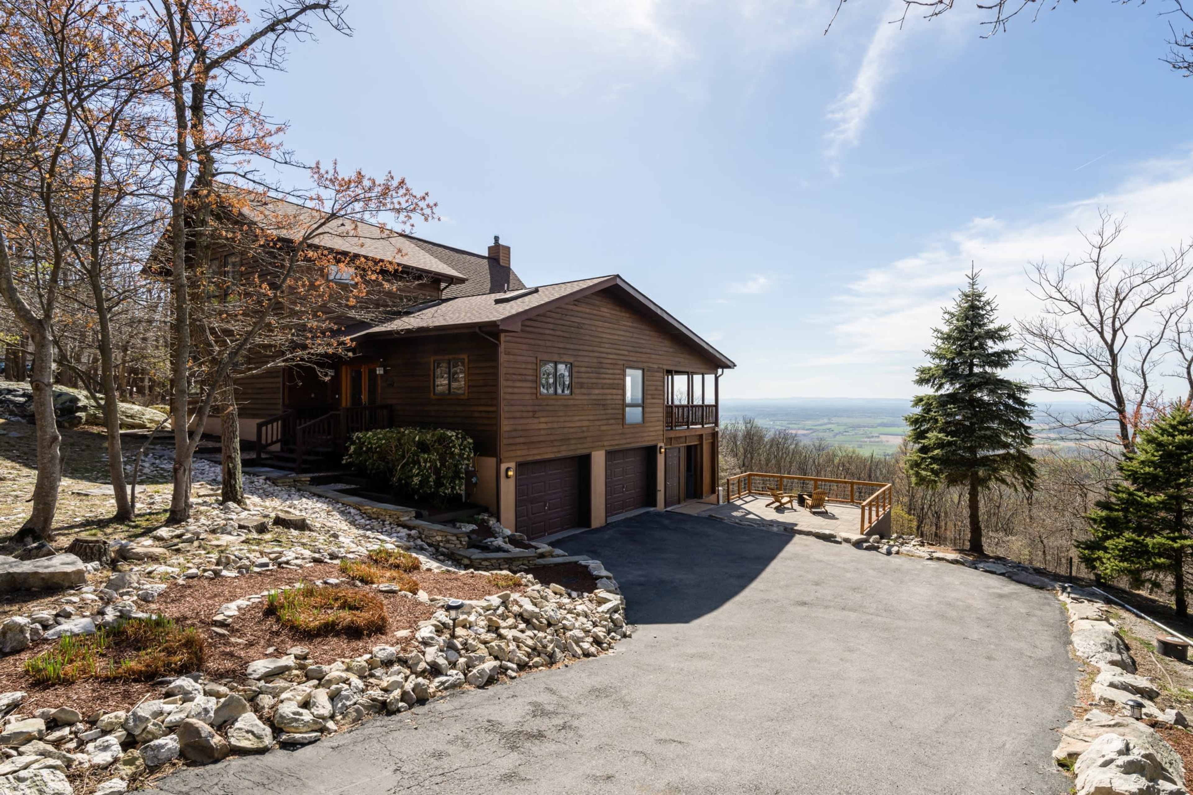 A large wooden house is situated on a hillside with a paved driveway and views of the valley below.