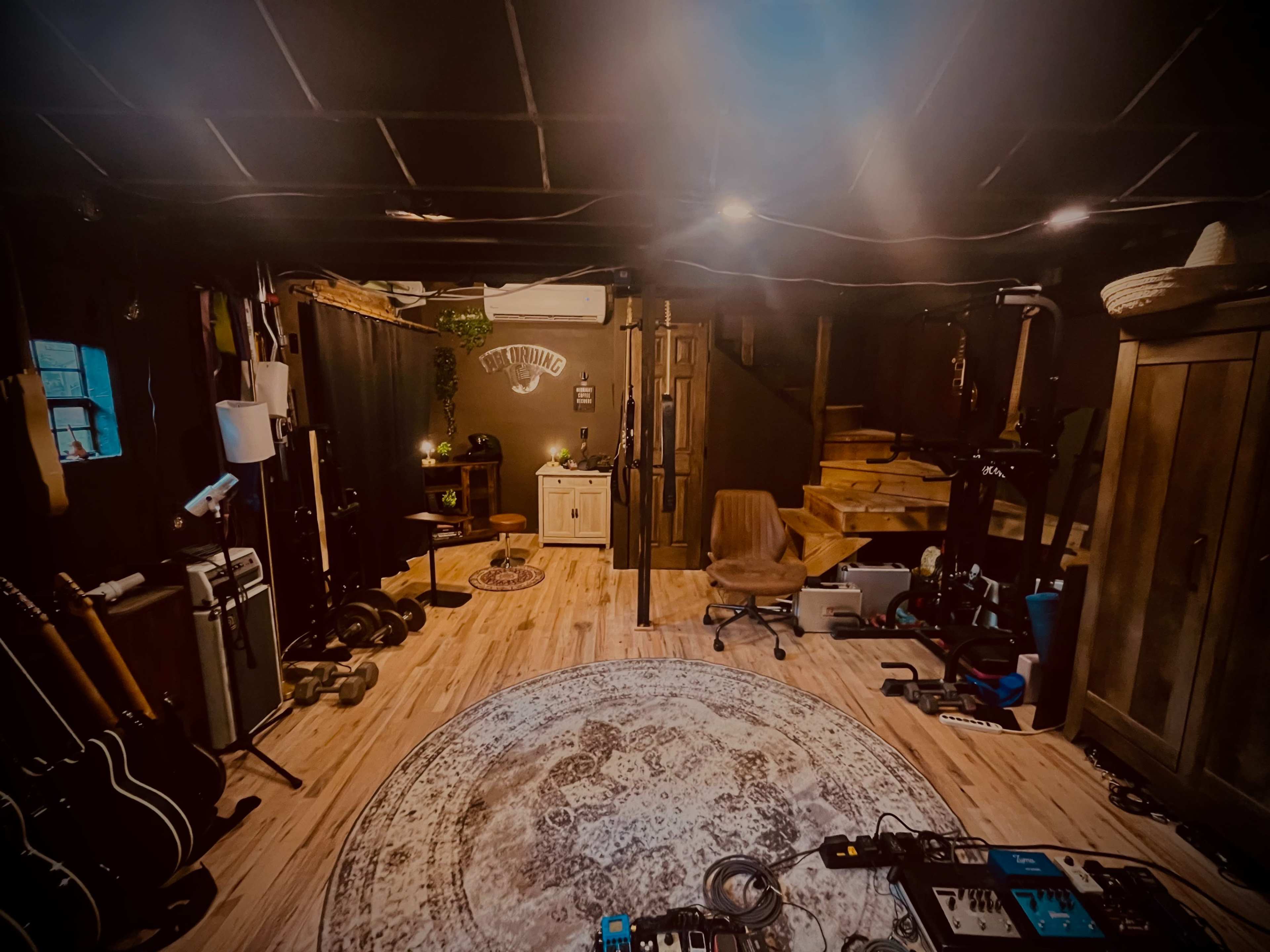The image shows a dimly lit basement studio with musical instruments, a rug in the center, and a staircase leading to another level.