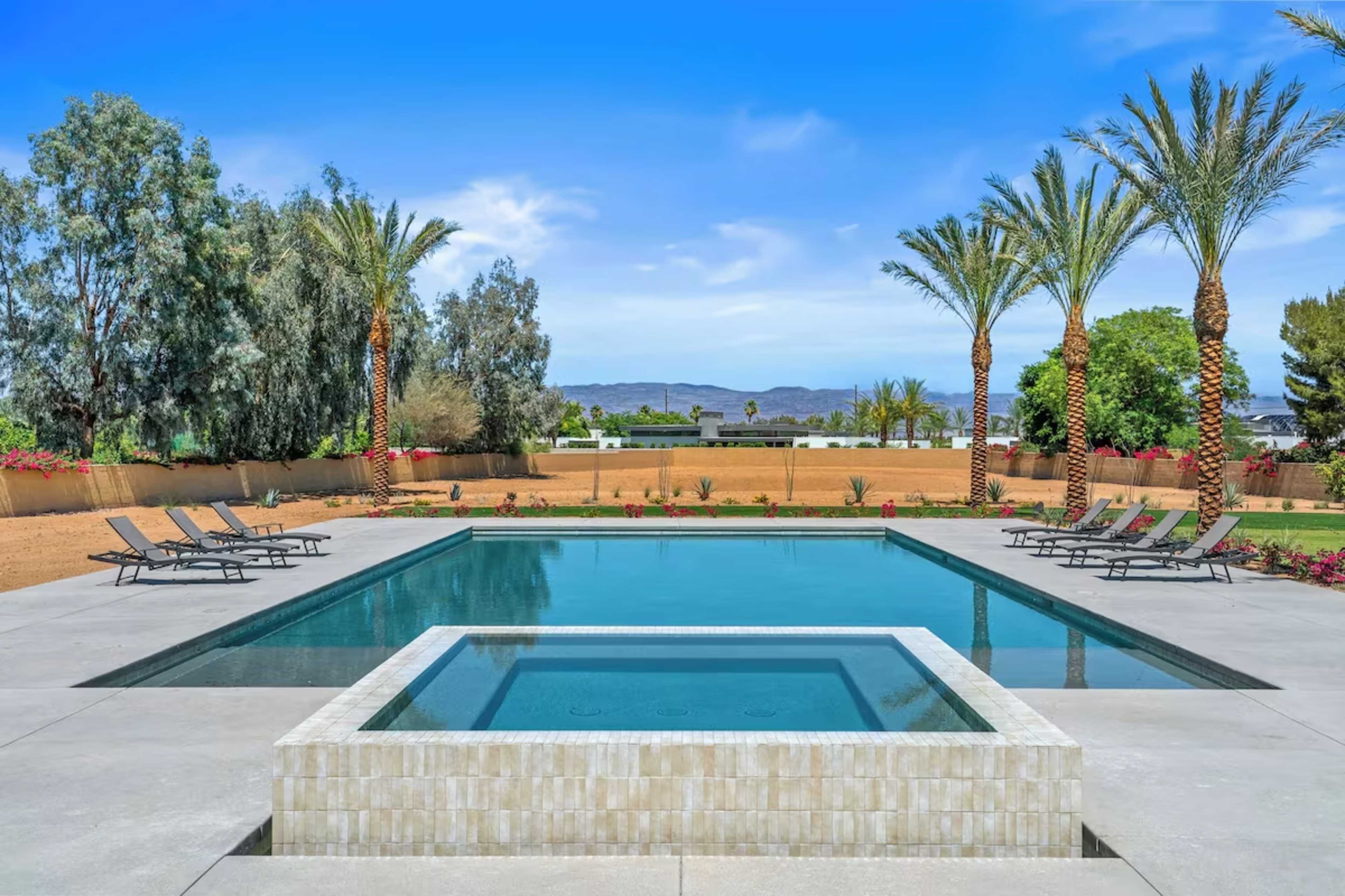 The image shows a rectangular swimming pool surrounded by lounge chairs and palm trees, with a scenic backdrop of a clear blue sky and distant mountains.