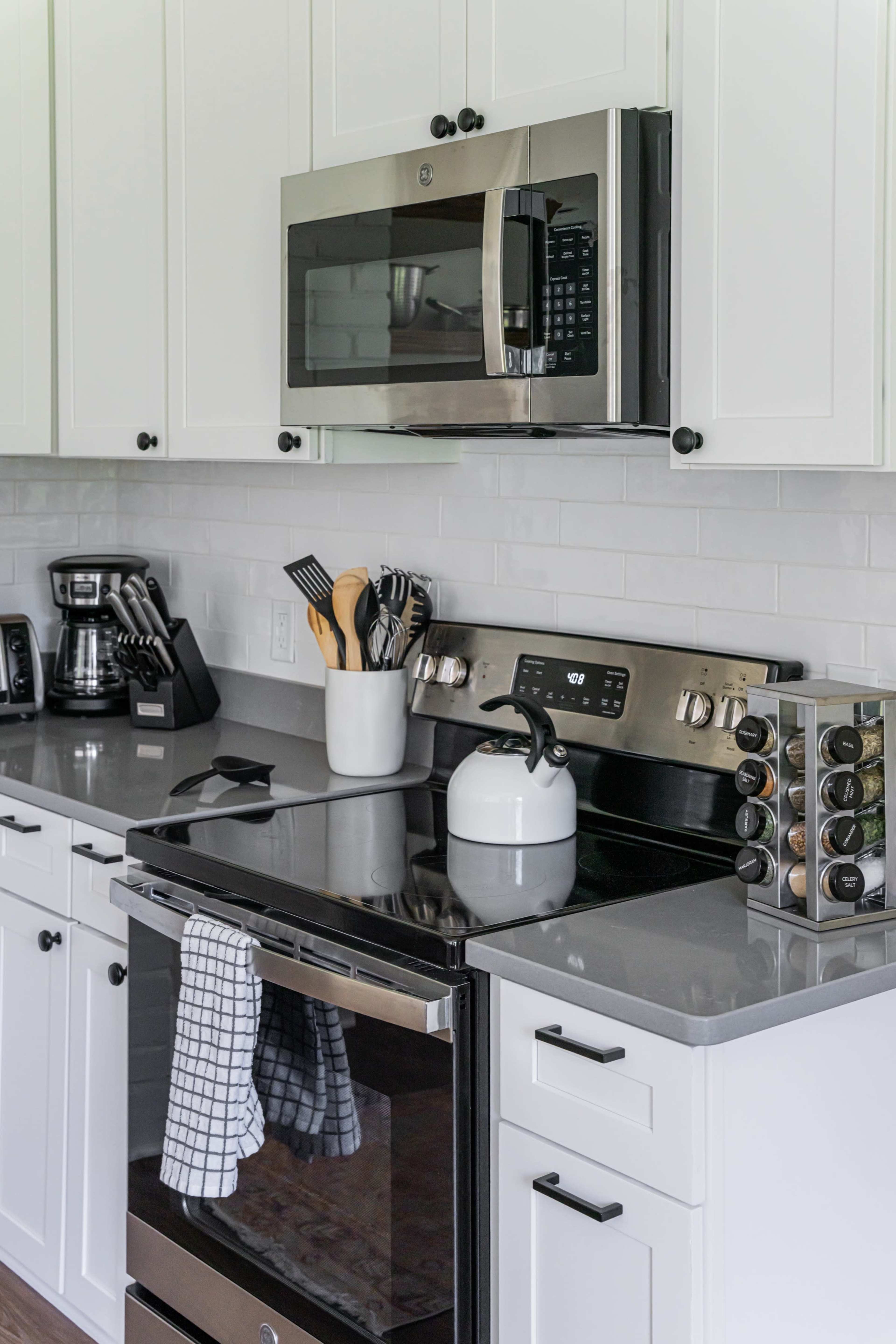 A modern kitchen features white cabinets, a gray countertop, an oven with a black glass door, a stainless steel microwave, and various cooking utensils neatly arranged.