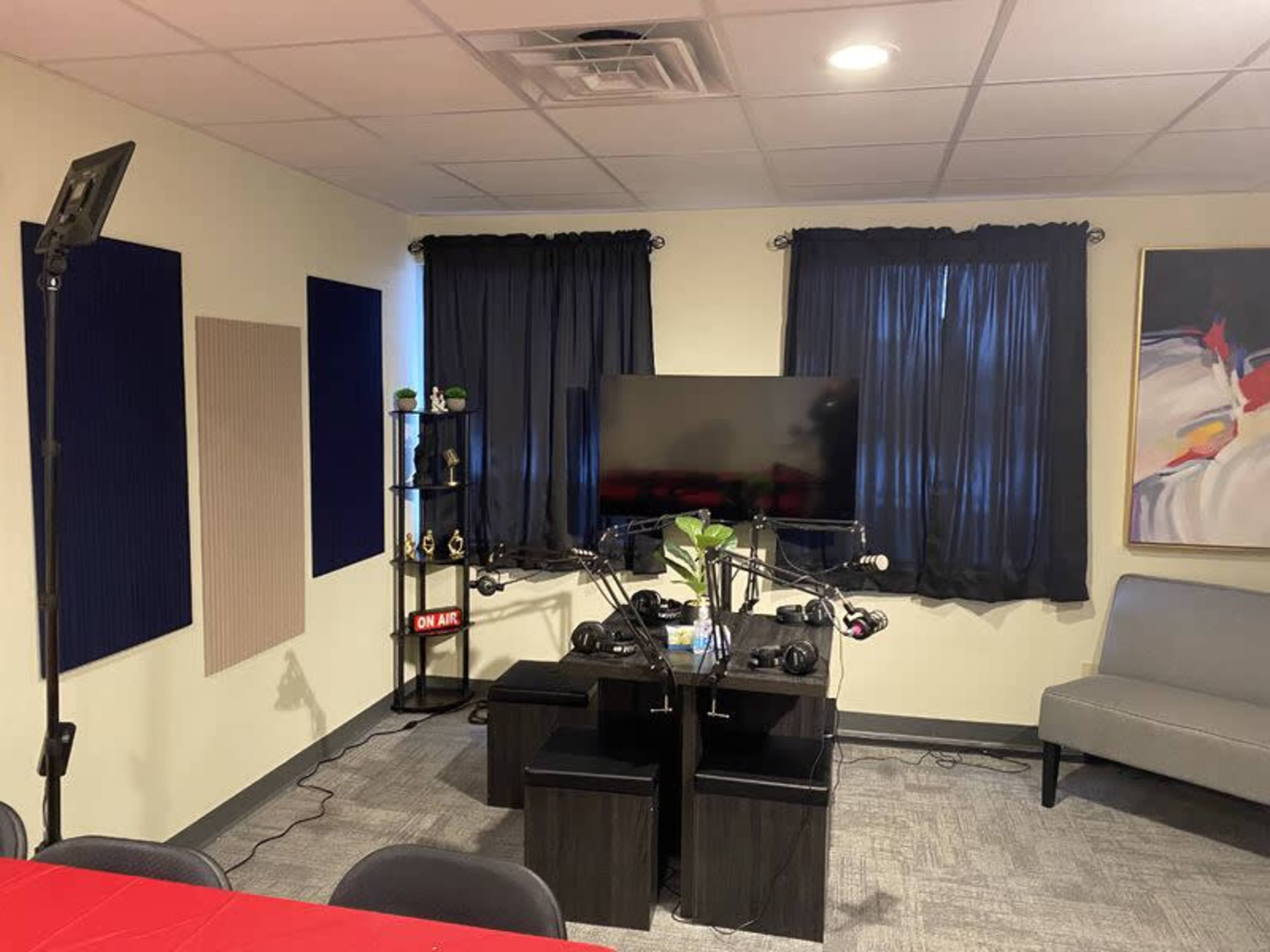 A room set up for recording or broadcasting, featuring a large TV, a table with microphones and headphones, and decorated with dark curtains and artwork.
