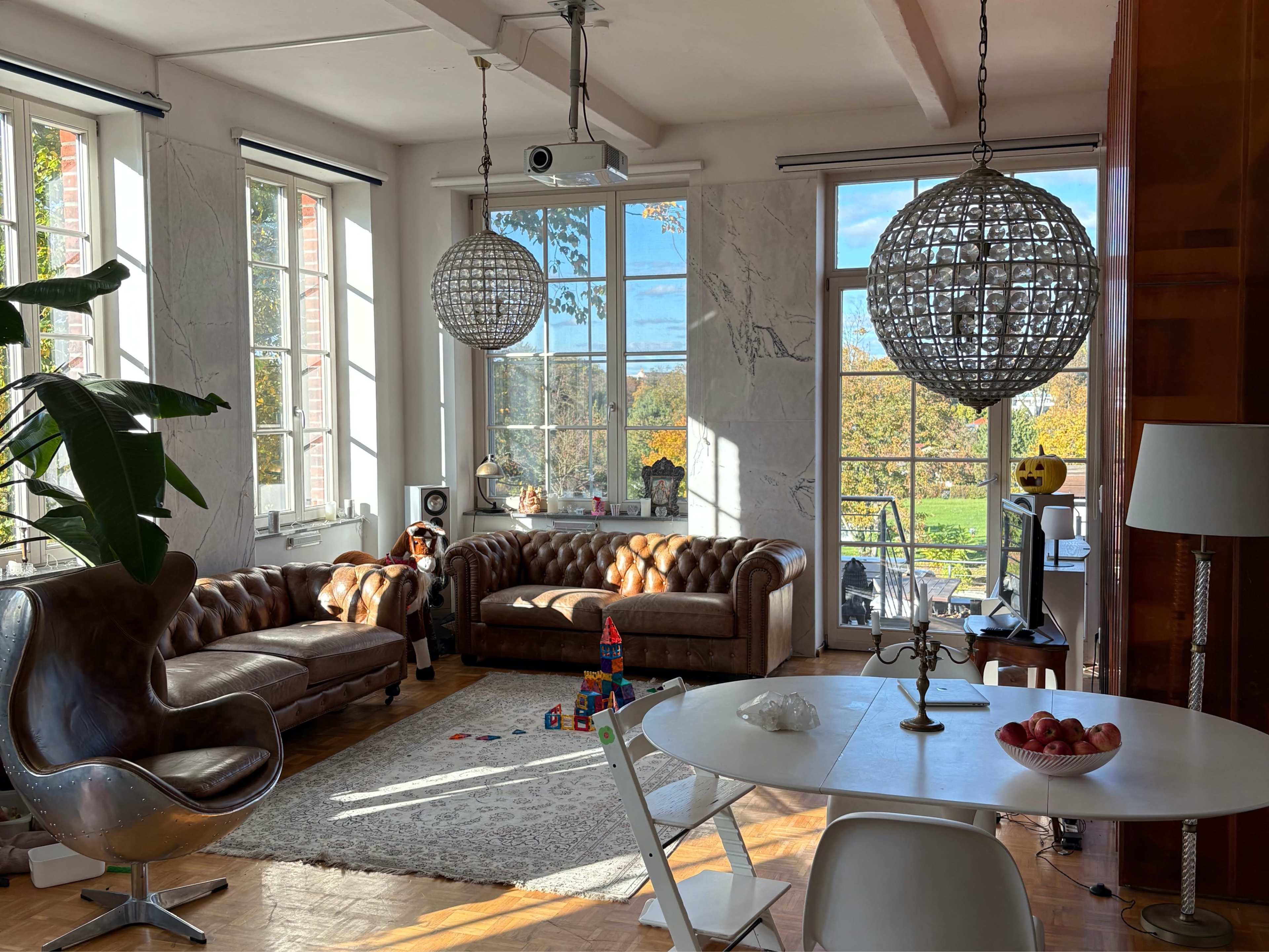A spacious living room features two brown leather sofas, a round dining table, and large windows overlooking a view with trees.