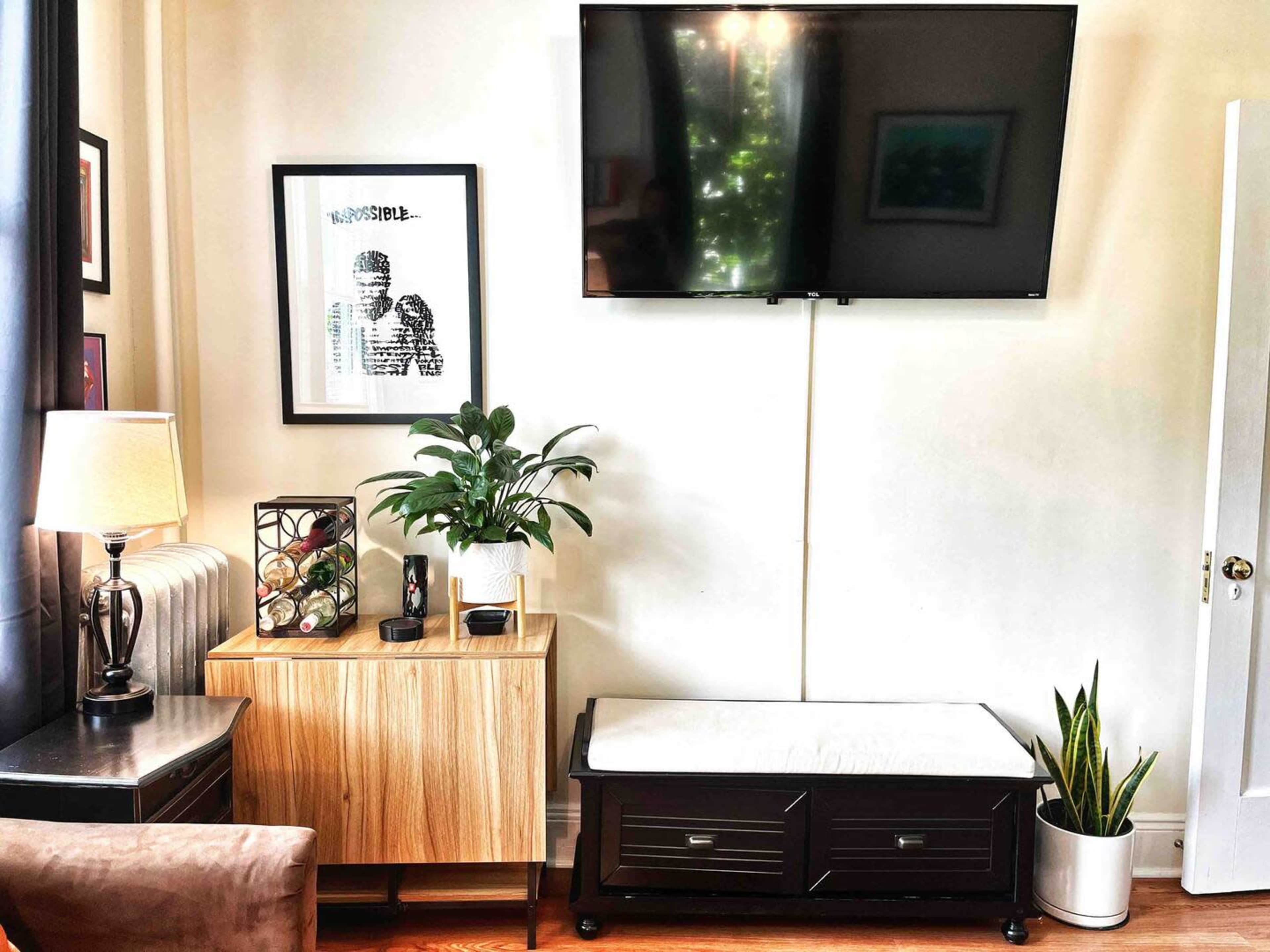 A living room setup featuring a television mounted on the wall, a light wood side table with a framed artwork, a lamp, and a potted plant beside a black storage bench and another potted plant in a corner.