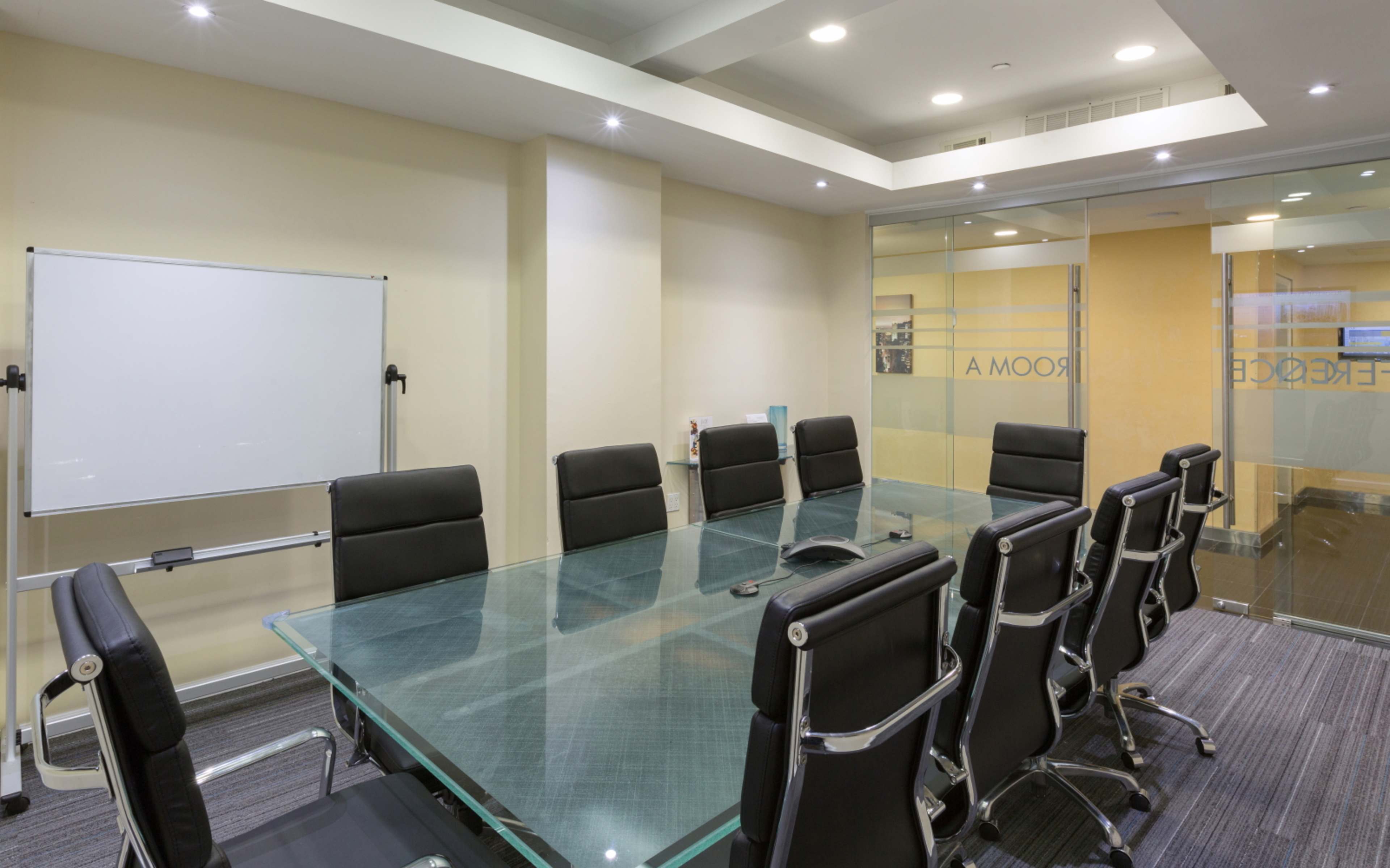 A modern conference room features a large glass table surrounded by black chairs and a whiteboard, with glass walls separating it from an adjacent space.