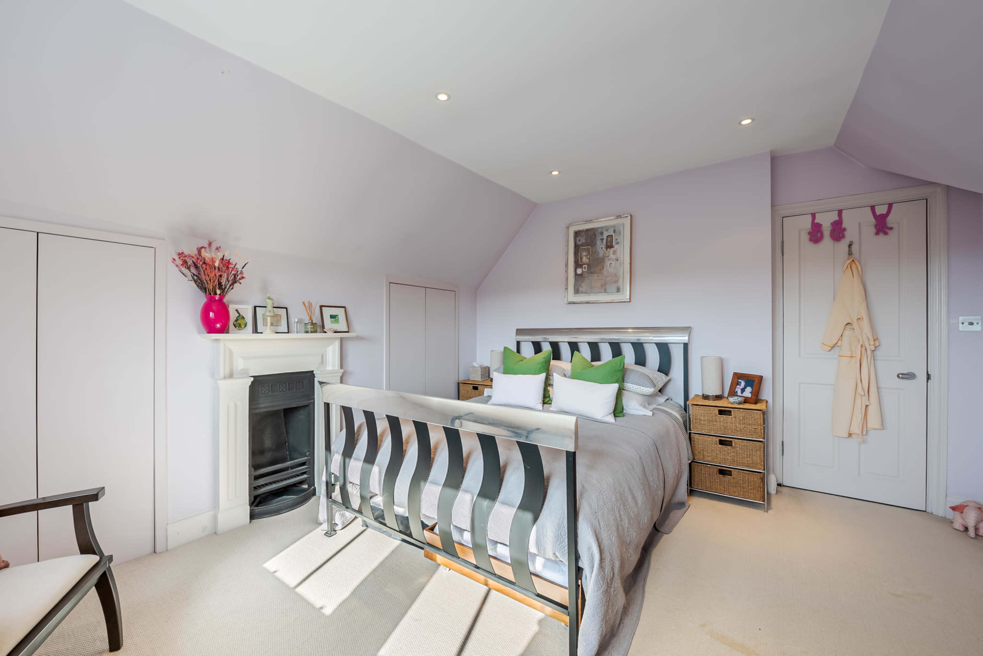 A light-colored bedroom features a bed with green and white pillows, a fireplace, and a chair beside a closet.