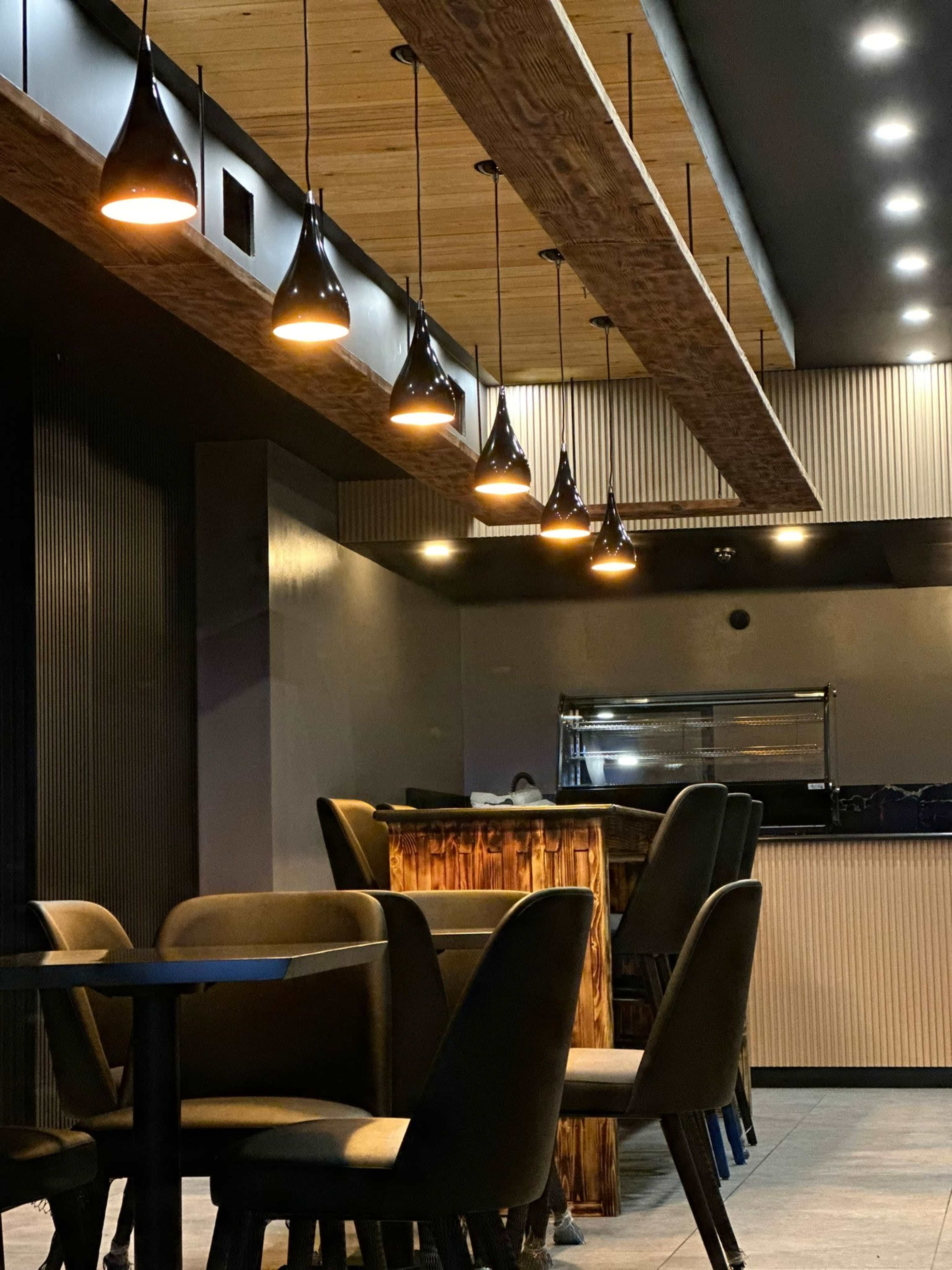 A modern restaurant interior with a dark color scheme, wooden accents, and pendant lights hanging above a bar area surrounded by chairs and tables.