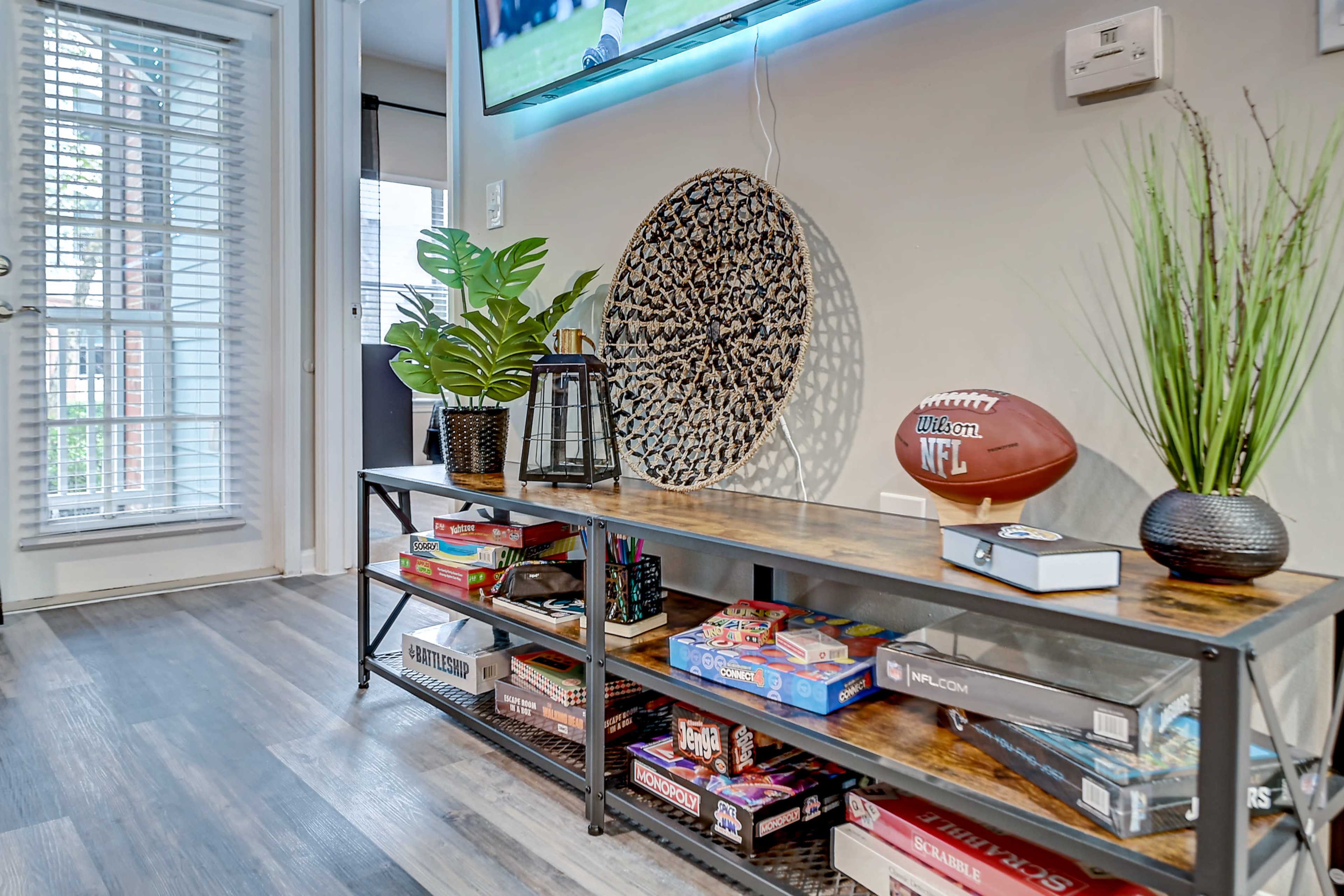 A living room features a console table with decorative plants, a football, and several board games displayed underneath a mounted television.