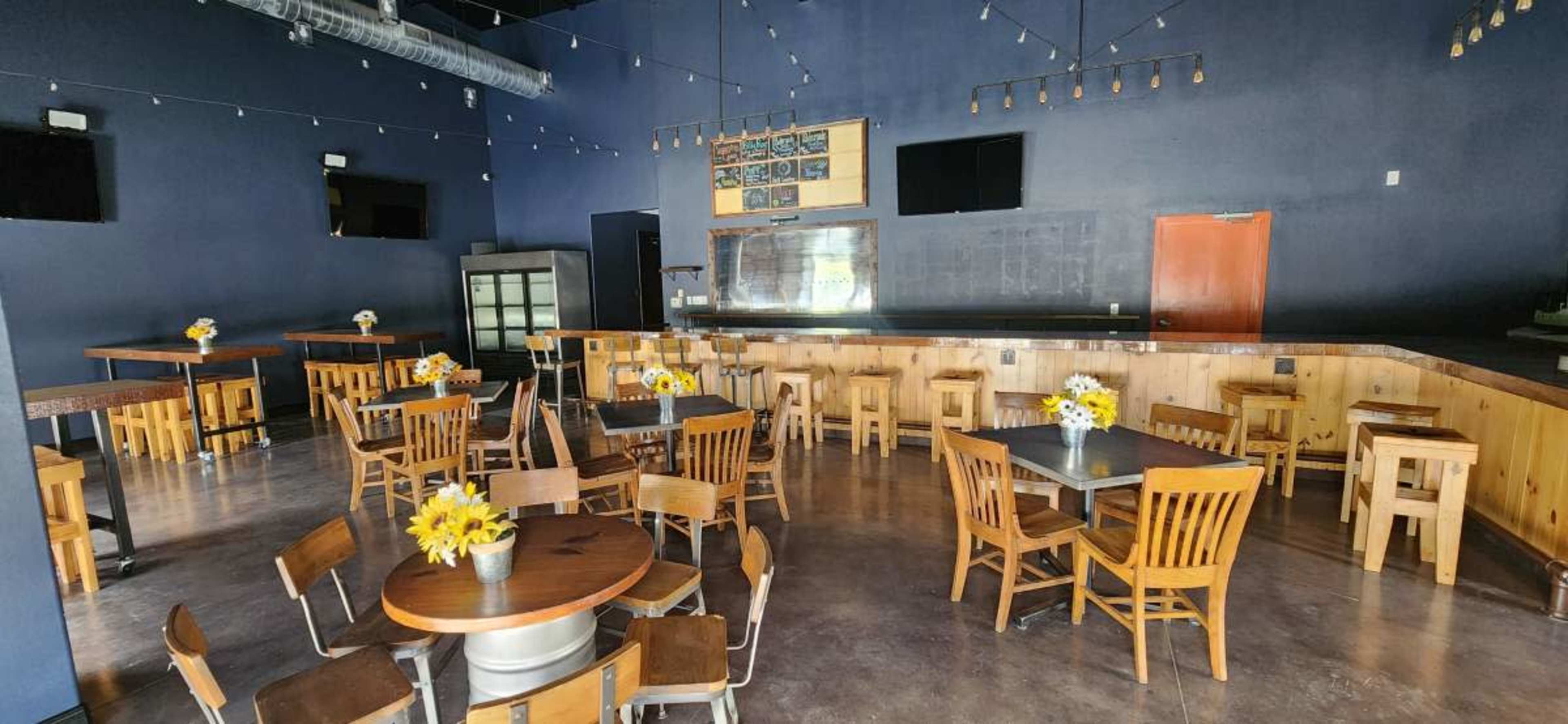 The image shows a spacious, dimly-lit restaurant interior with wooden tables and chairs arranged around a bar area, decorated with small flower centerpieces.