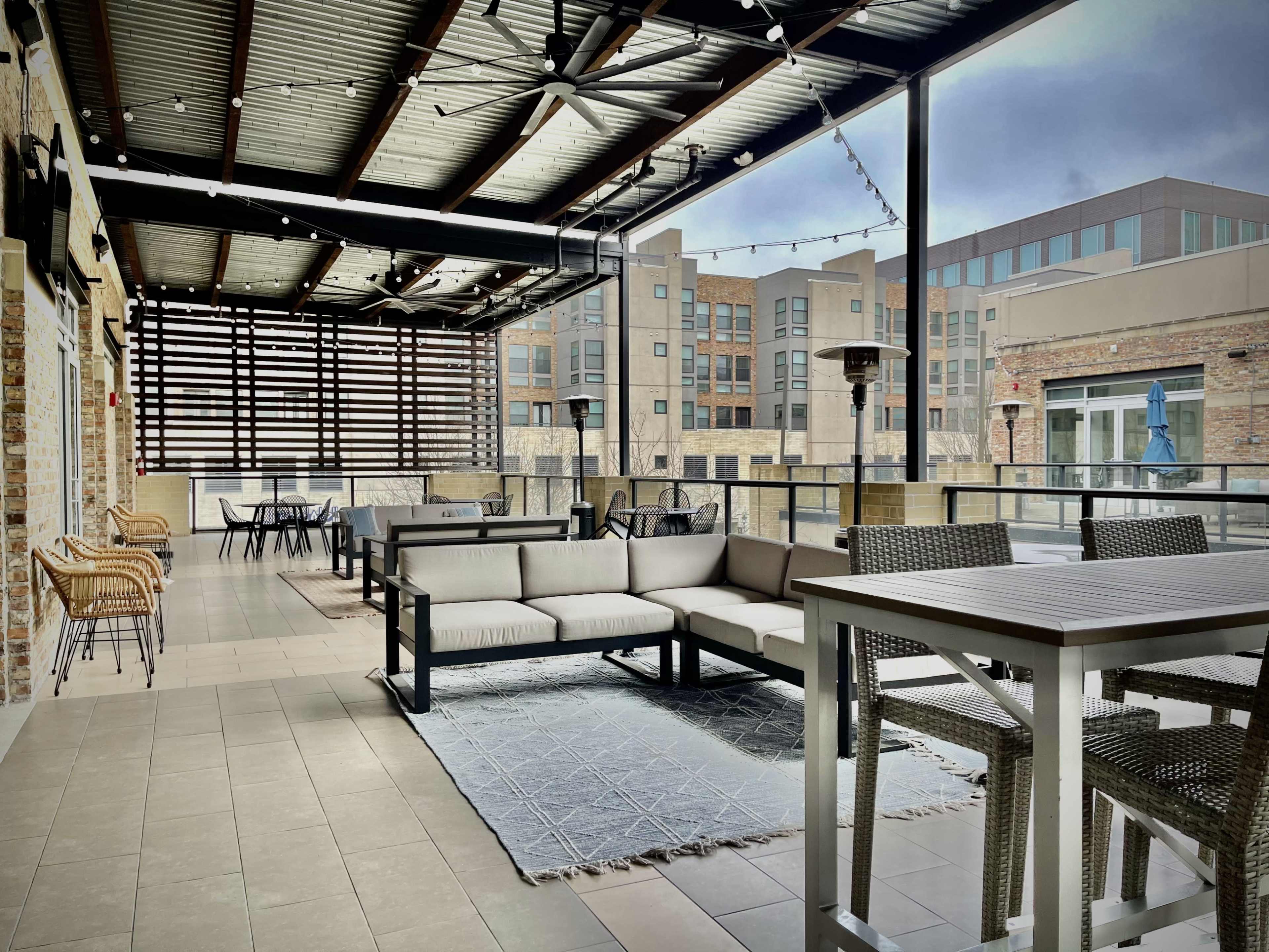 A spacious outdoor terrace with modern seating arrangements, a table, and overhead lighting, overlooking a multi-story building complex.