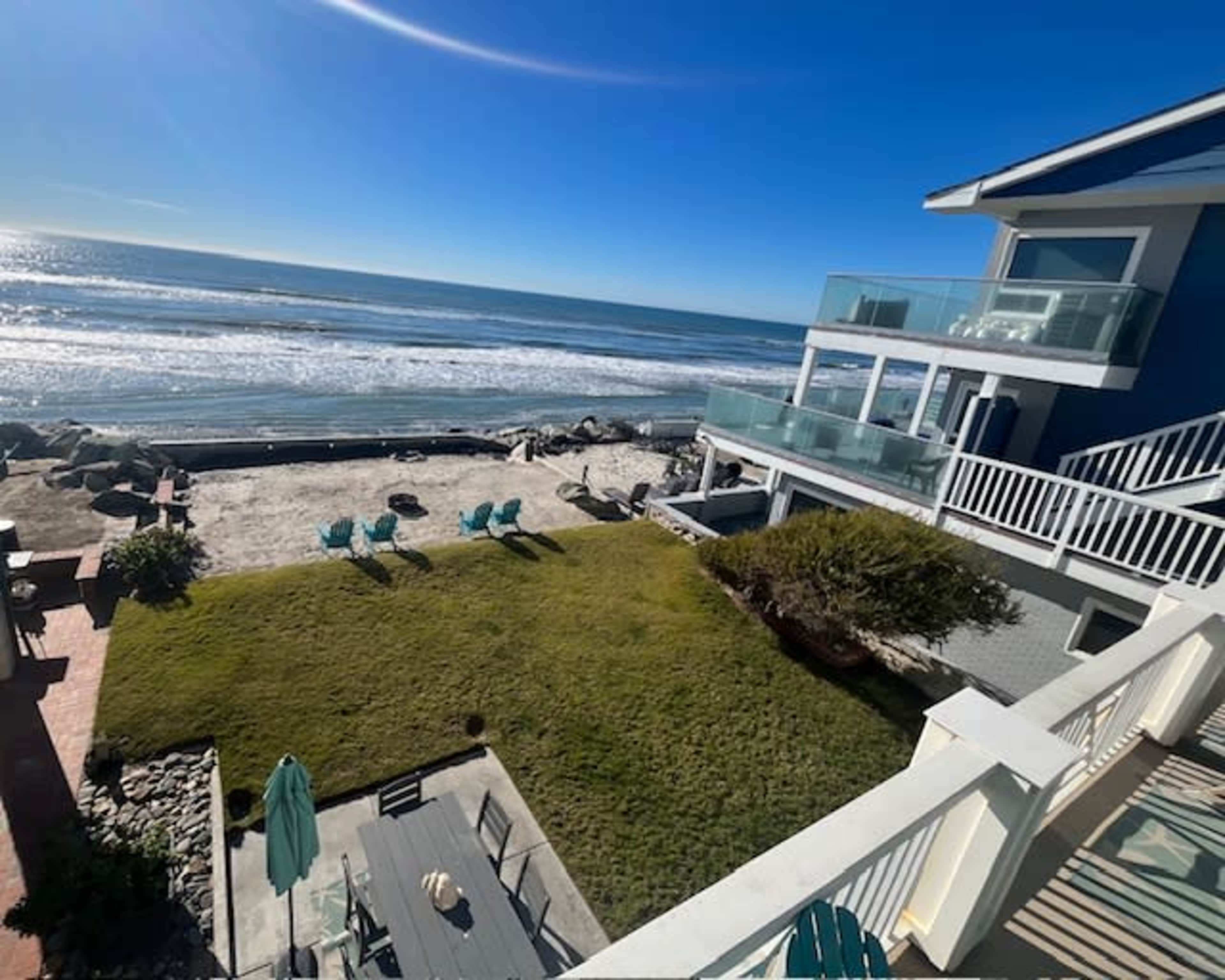 The image shows a beachfront property with green lawn furniture, overlooking the ocean and a sandy beach.