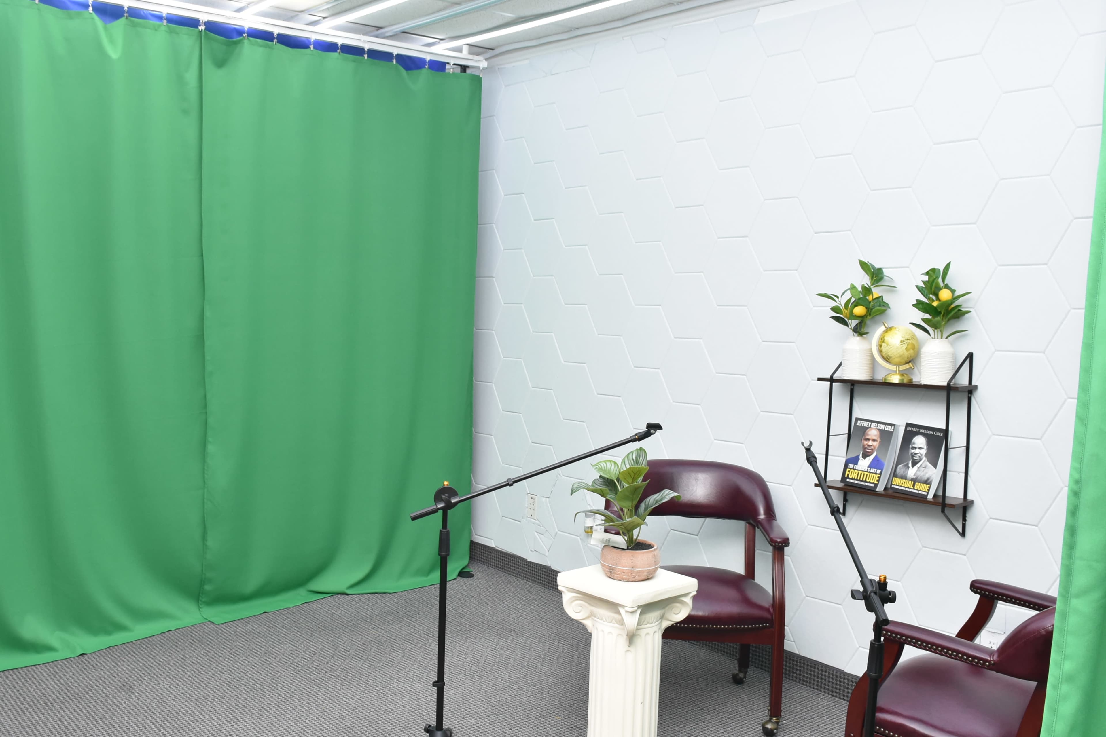 The image shows a small studio setup with two chairs, a small table, and green curtains on either side of the space, suggesting it is intended for filming or interviews.