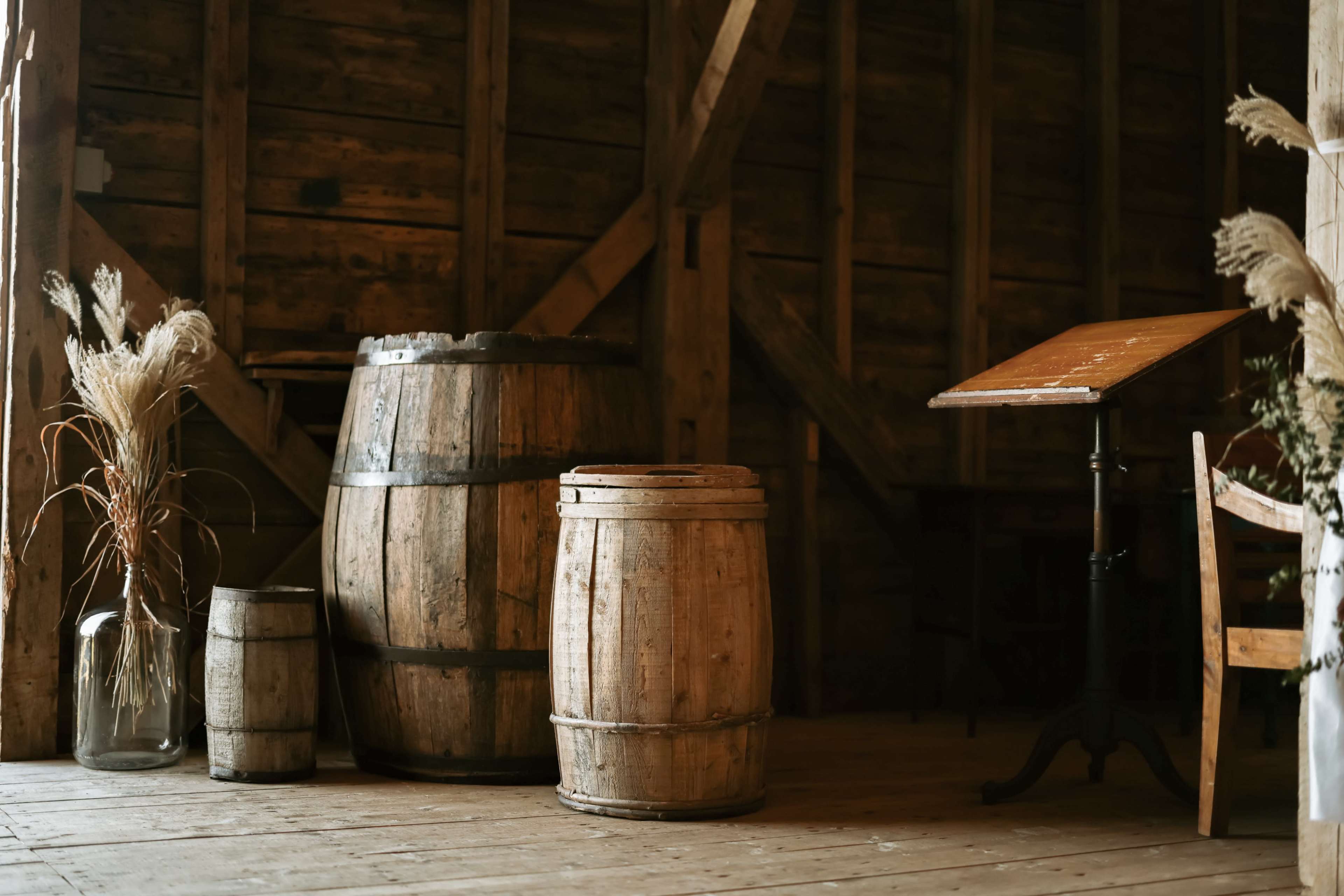 The image shows a rustic interior featuring wooden barrels, a small table, and a glass vase with dry grass arrangements.