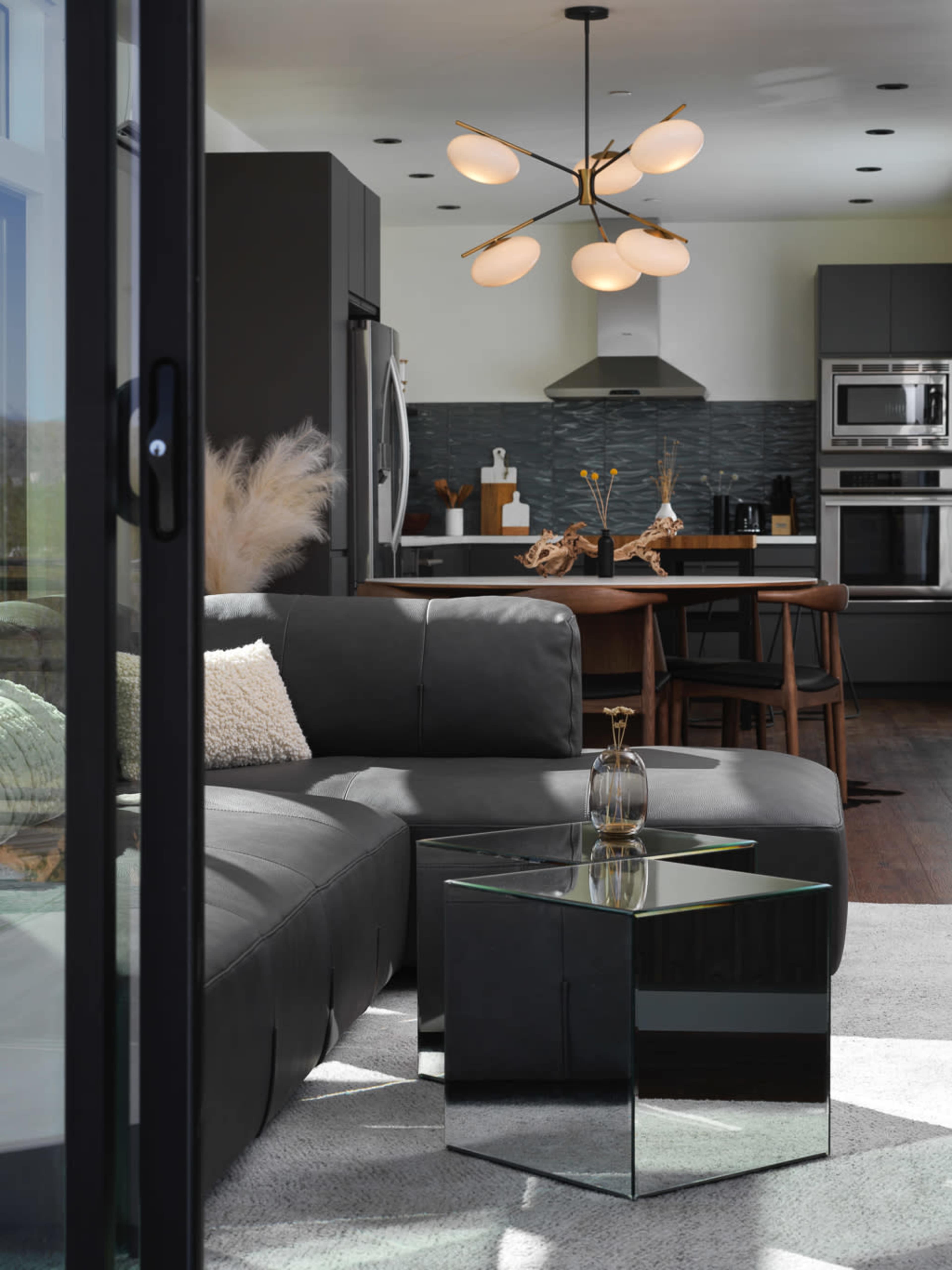The image shows a modern living room with a gray sectional sofa, a glass coffee table, and a stylish light fixture, leading into a kitchen area with sleek appliances.