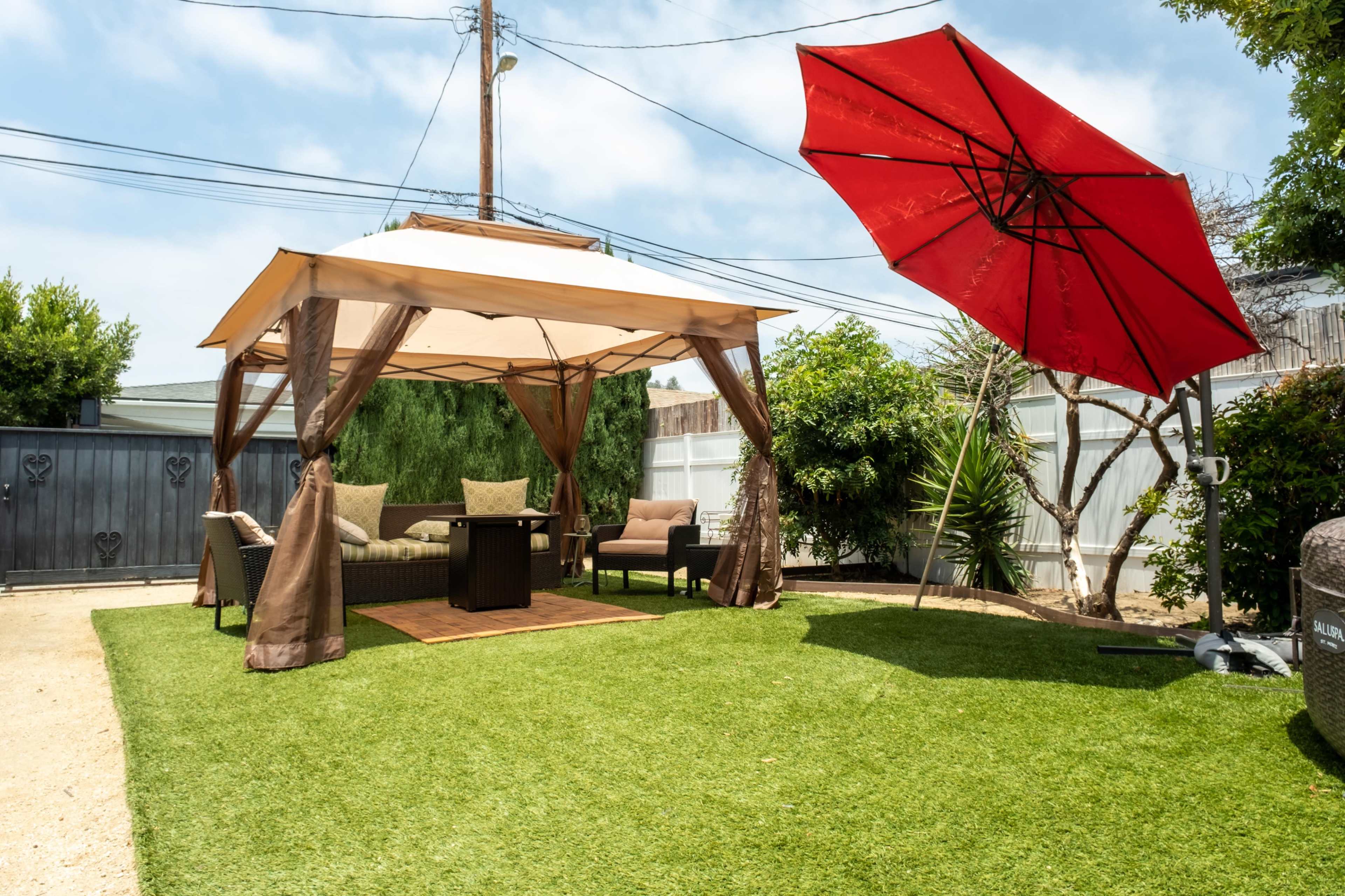 A shaded seating area with a gazebo and a large red umbrella is set up on green artificial grass in a backyard.