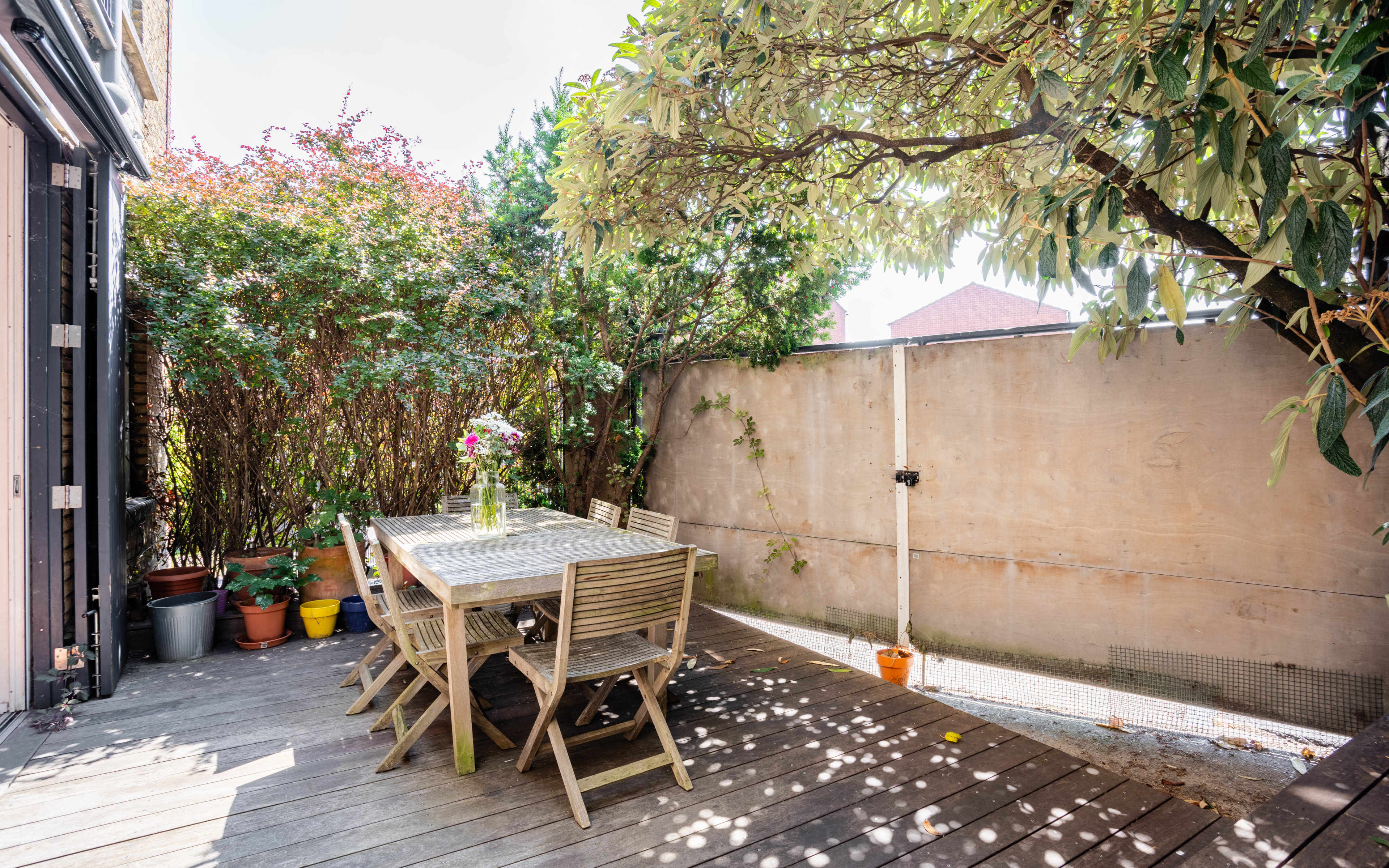 A wooden outdoor dining table with chairs is set up on a deck surrounded by greenery and potted plants.