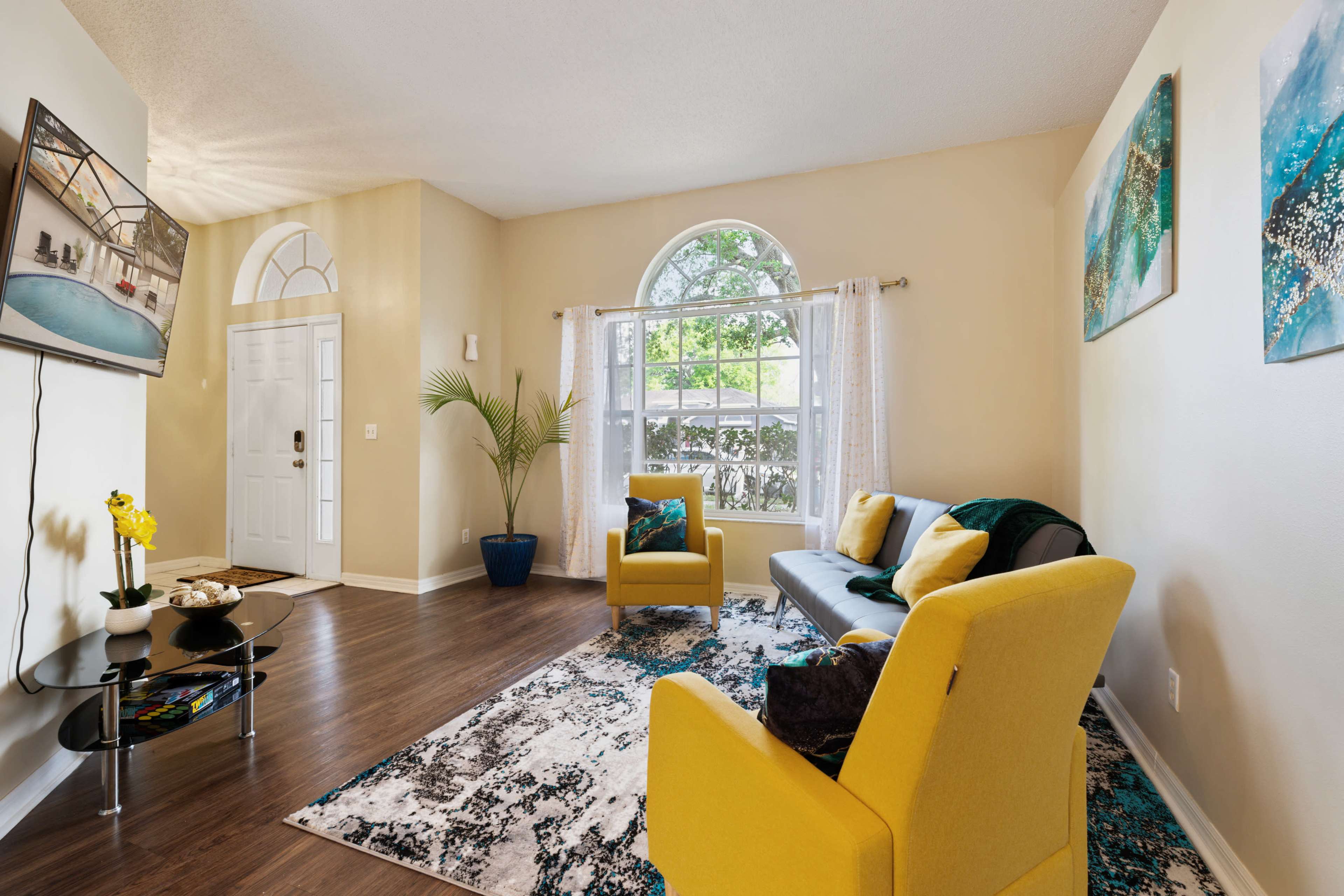 A living room features a gray sofa, two yellow chairs, and a large window with white curtains, surrounded by decorative artwork and a potted plant.