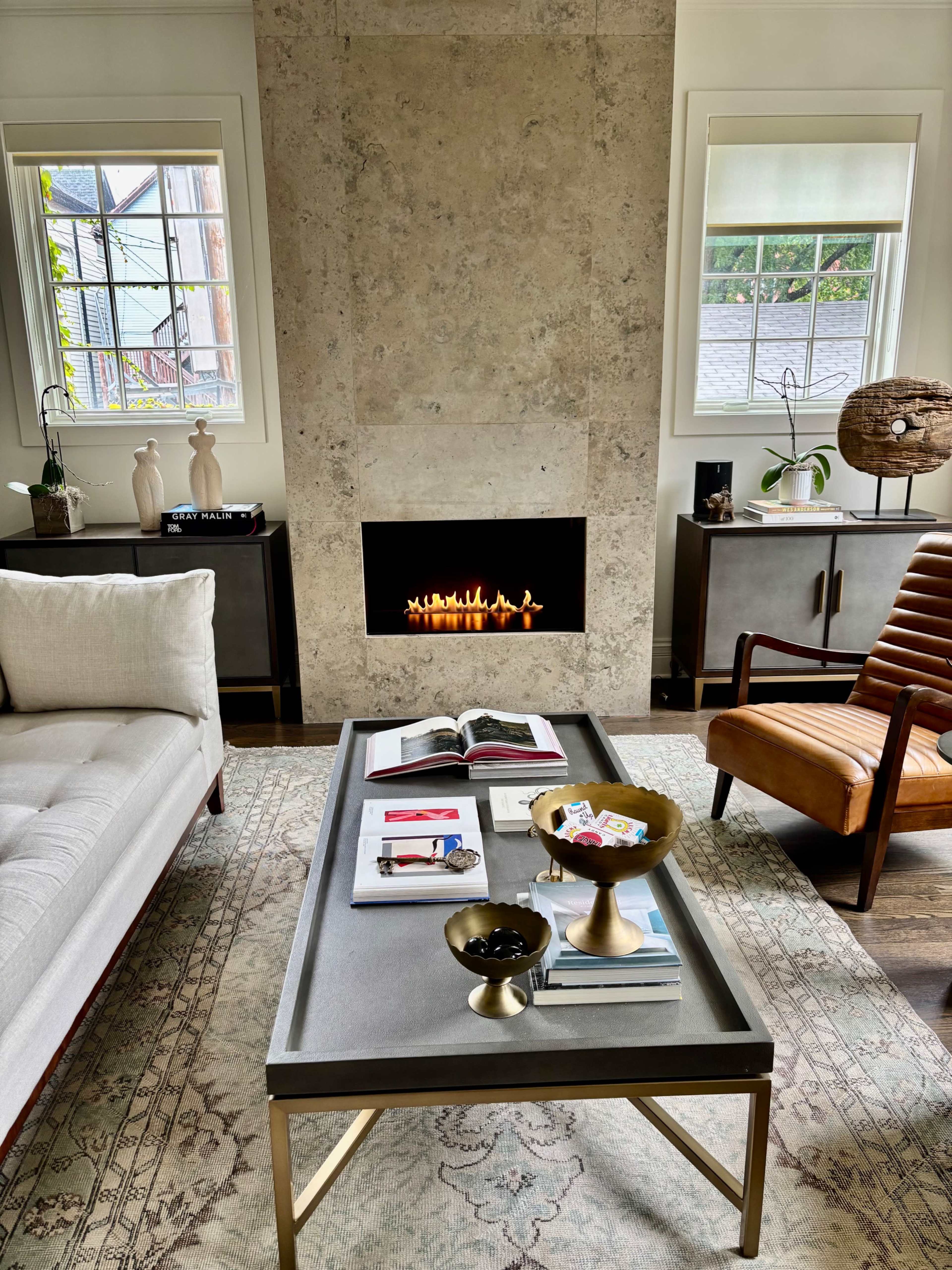 The image shows a cozy living room with a beige sofa, a wooden armchair, a low coffee table displaying books and decorative bowls, and a modern fireplace.