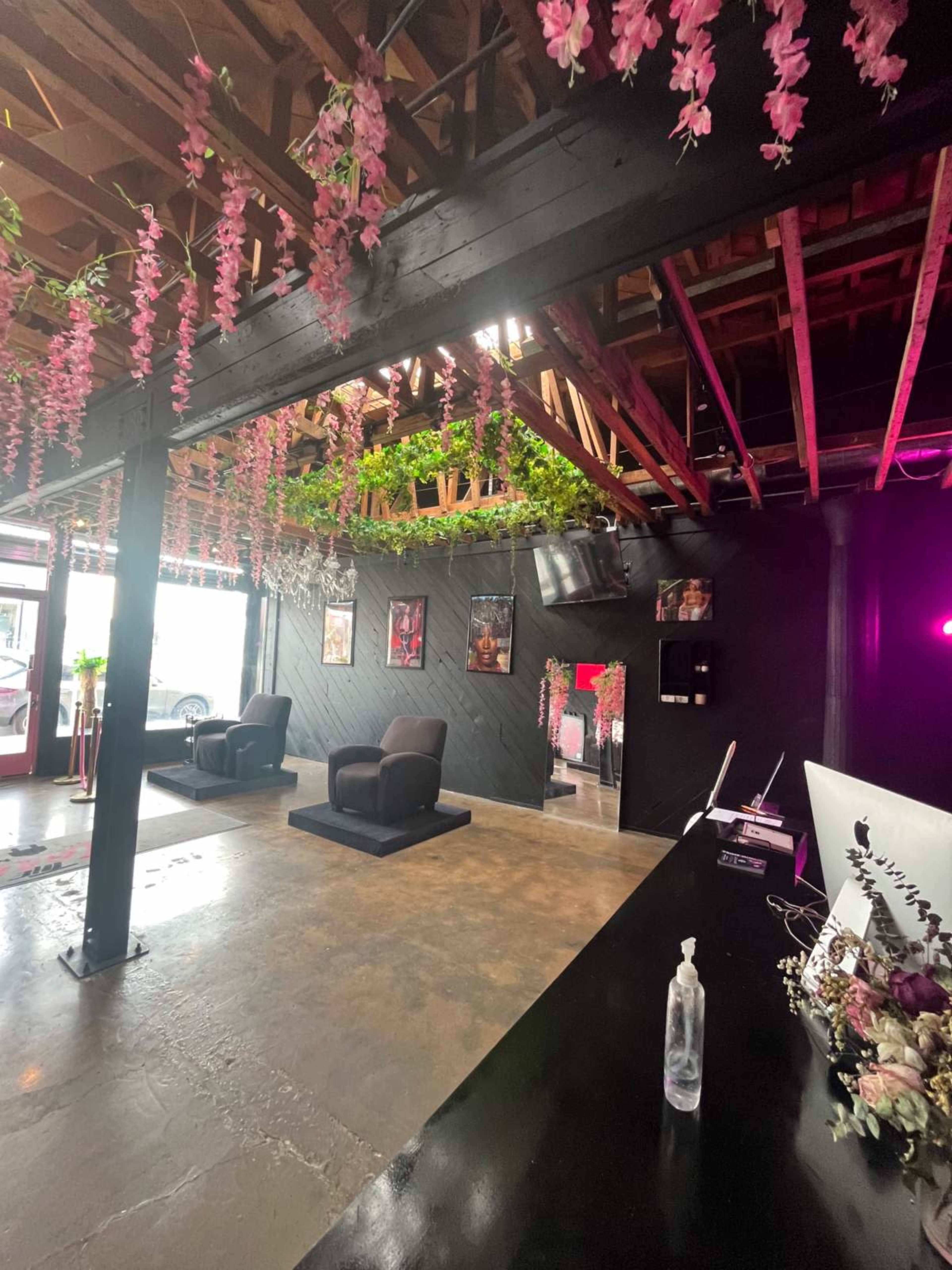 The interior of a salon features black walls, two chairs, and pink flowers hanging from the ceiling beams.