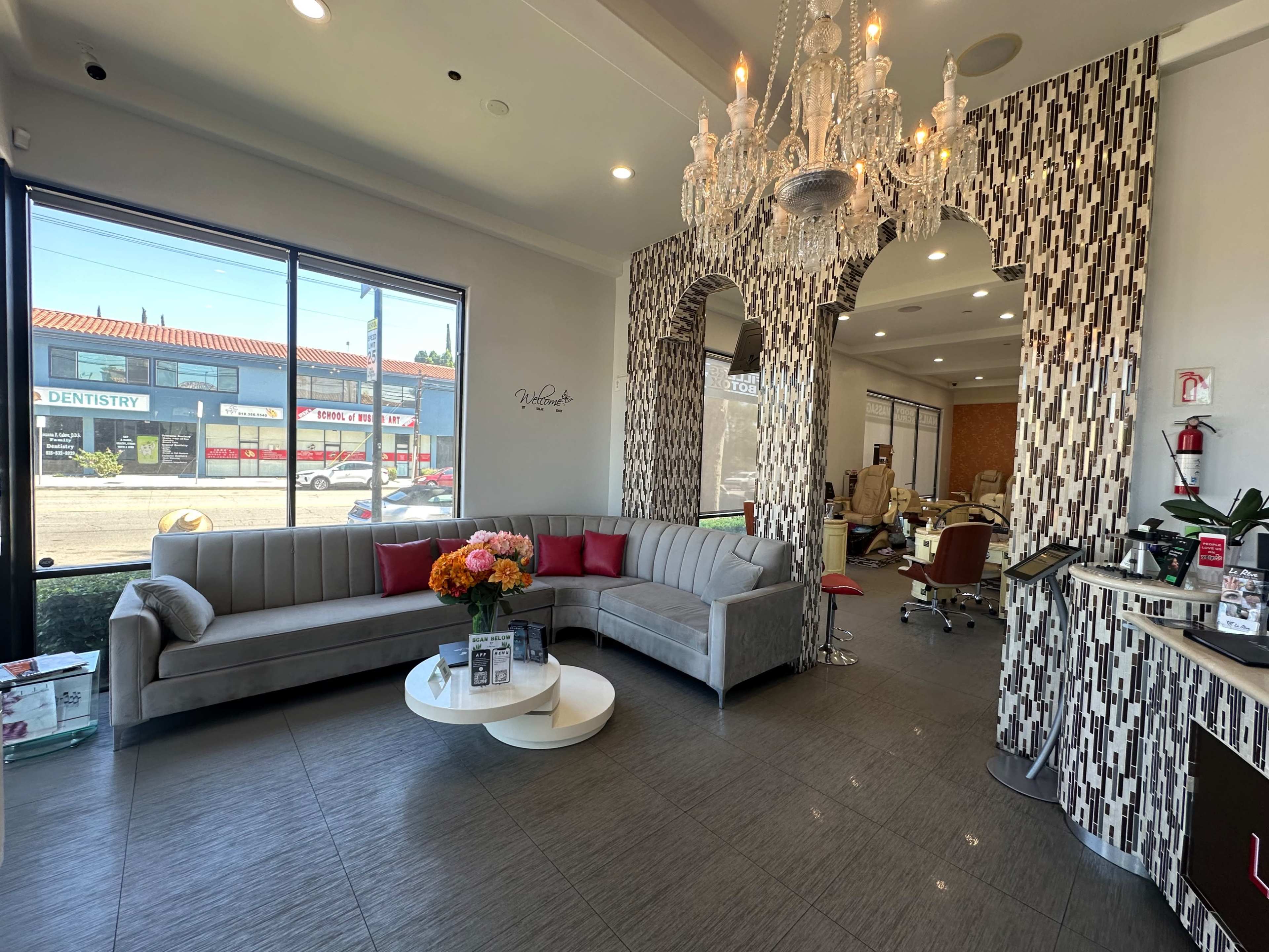 The image shows a modern salon interior featuring a plush gray sofa, a chandelier, and decorative wall patterns, with large windows viewing the street outside.